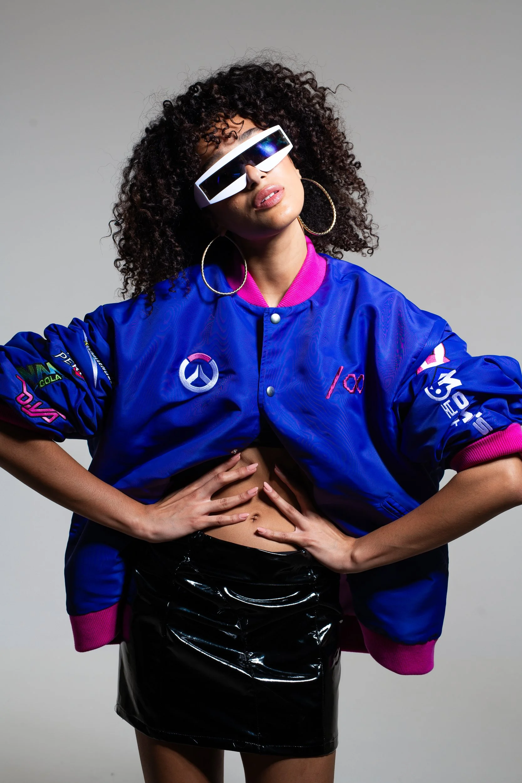 A woman with curly hair wearing futuristic sunglasses, a blue bomber jacket with pink accents and various patches, and a shiny black skirt.