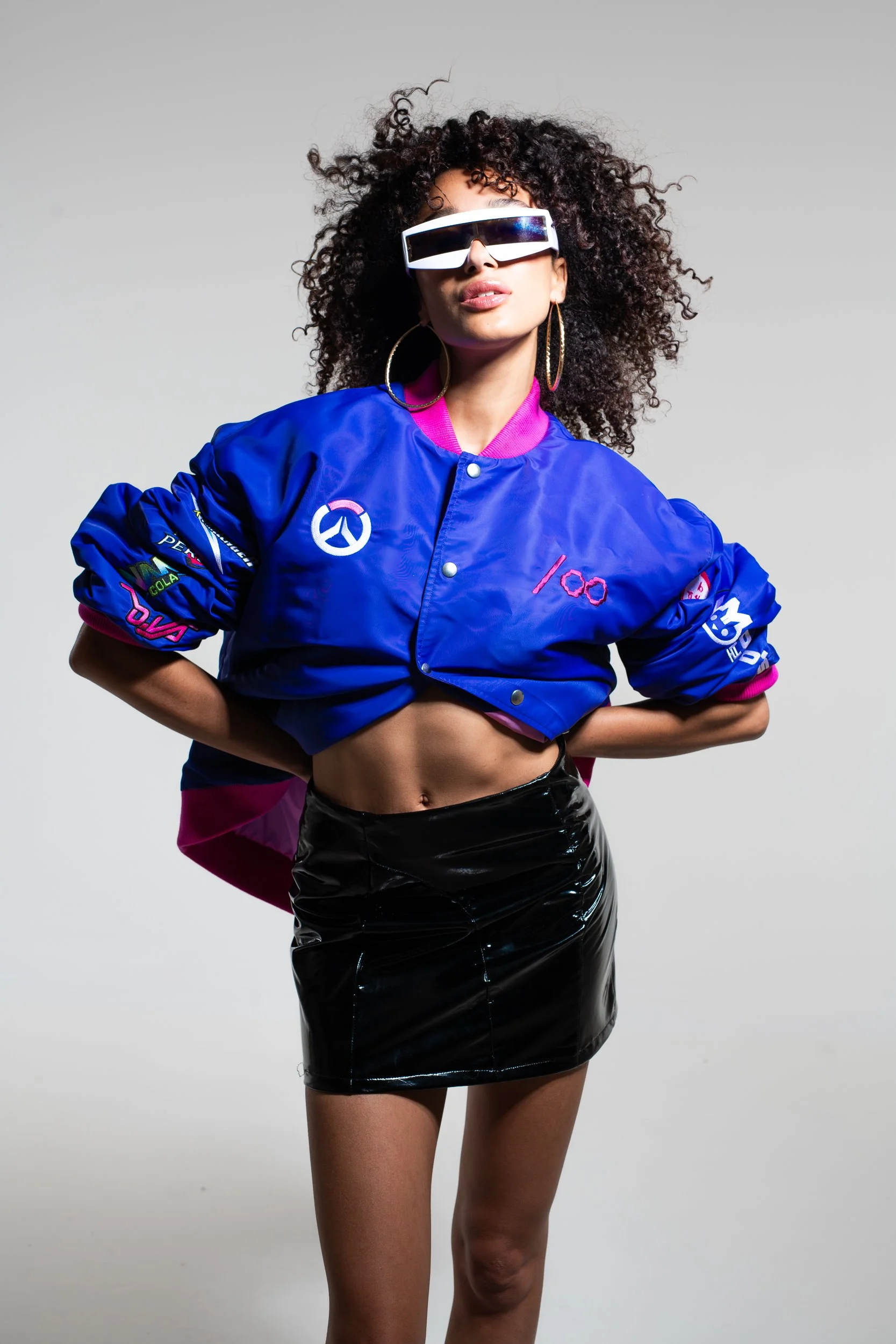 A woman with curly hair wearing large futuristic sunglasses, a blue and pink varsity jacket with patches, and a shiny black vinyl skirt.