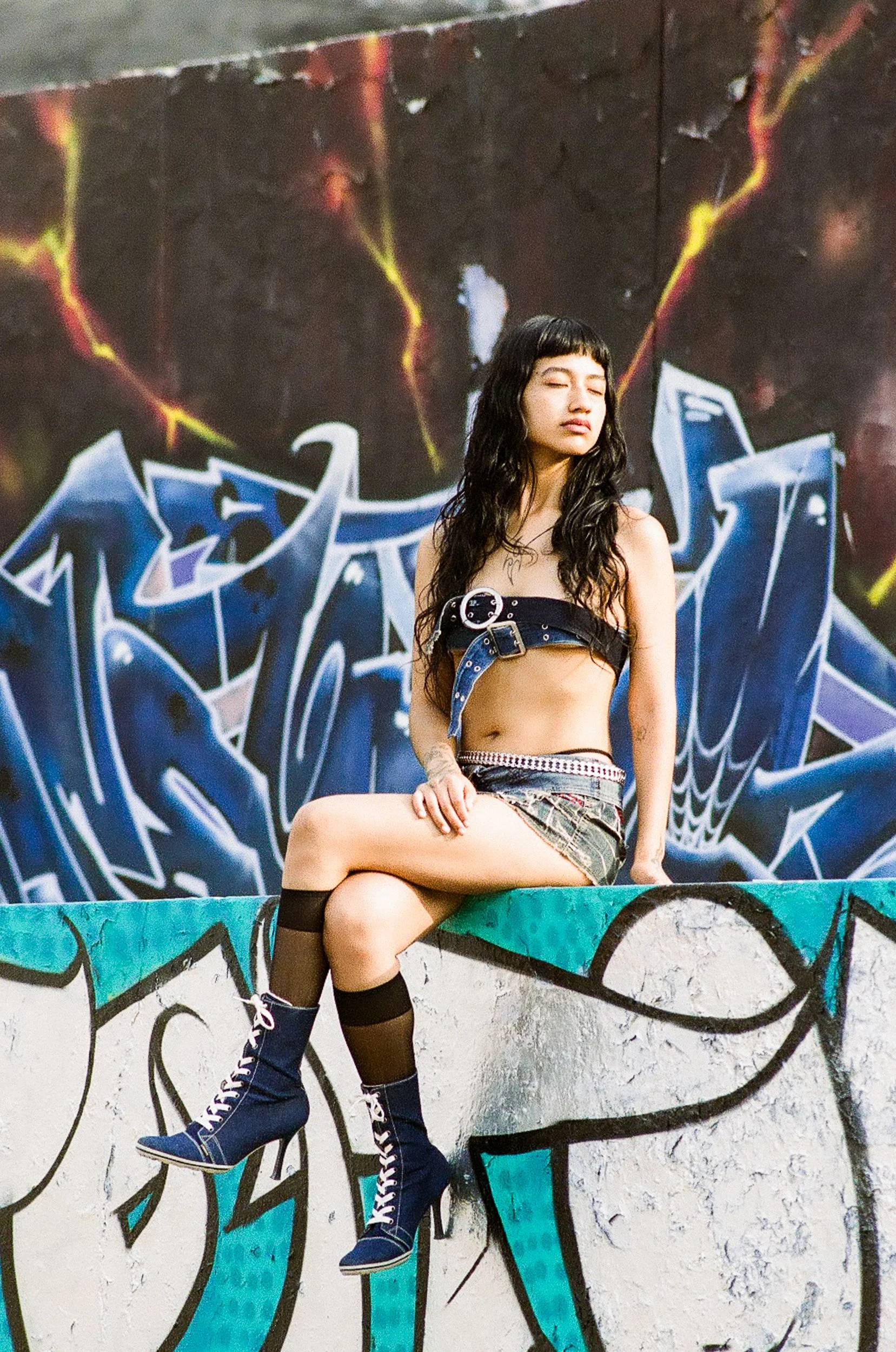 A young woman with long black curly hair, wearing a black strapless top, a short patterned skirt, and navy blue high-top sneakers, sitting on a colorful graffiti-covered ledge with her eyes closed, in front of a dark graffiti mural with vibrant, abst