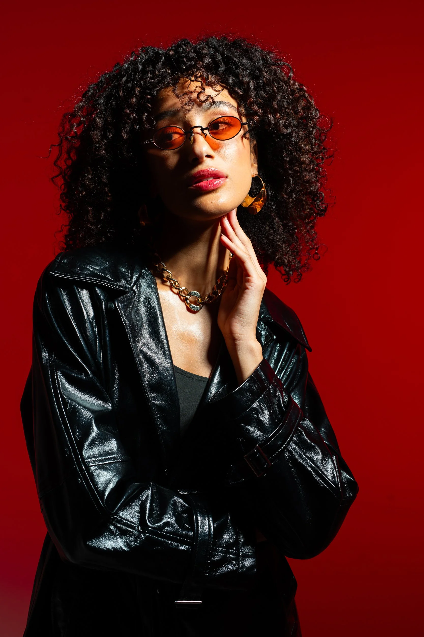 A woman with curly black hair wearing orange-tinted sunglasses, gold earrings, a chunky gold necklace, and a black leather jacket against a red background.