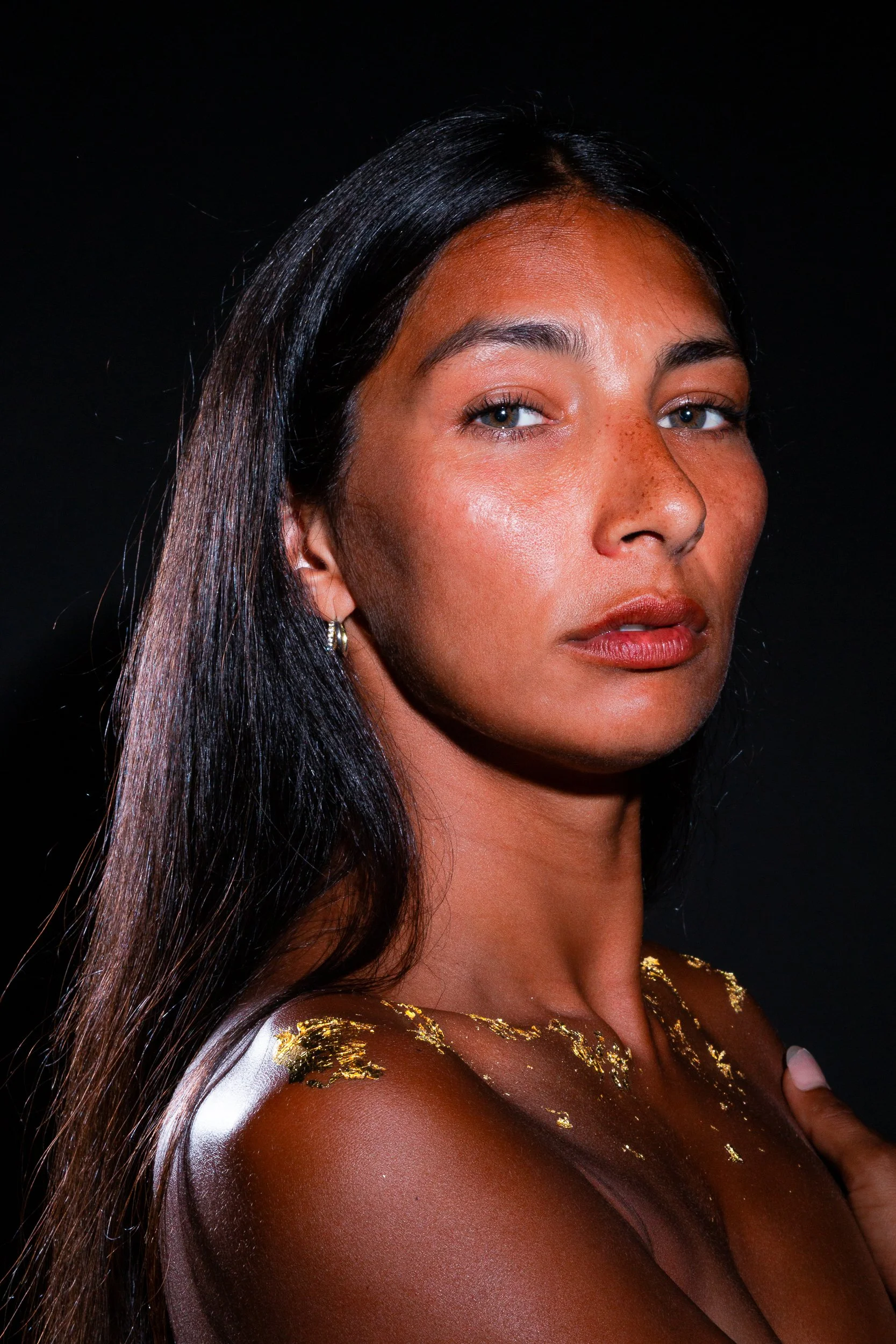 A close-up of a woman's face and upper shoulder, with long dark hair and makeup, against a black background.