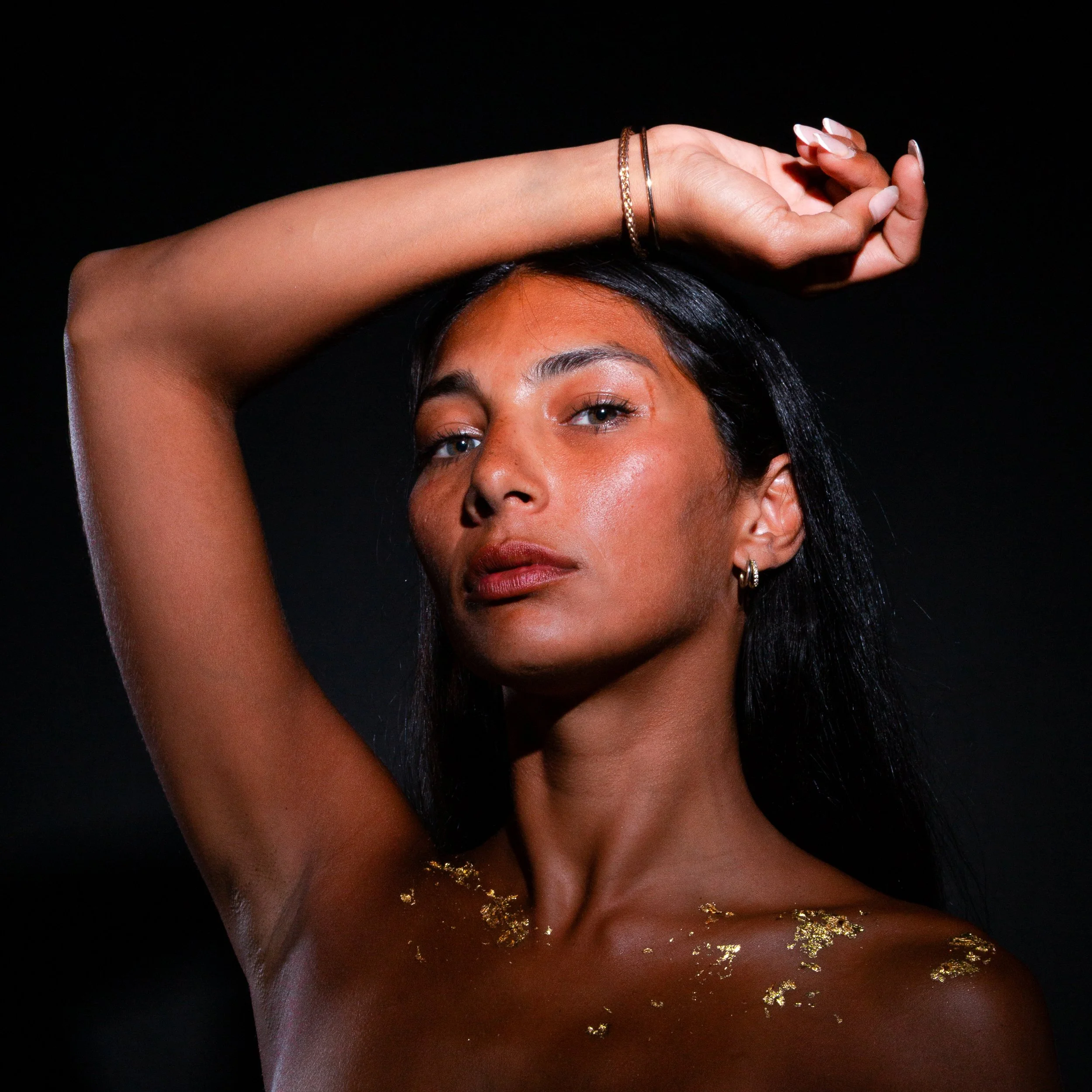 Close-up of a woman with gold glitter on her neck, wearing jewelry, with one arm raised above her head against a black background.