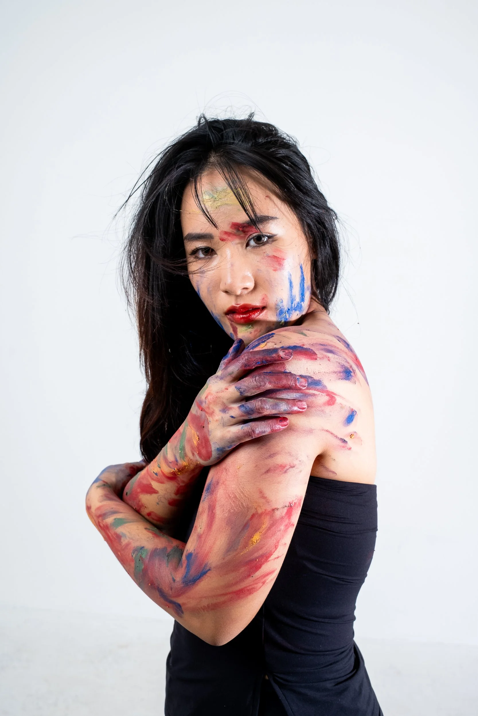 A woman with black hair covered in multicolored paint, standing against a plain white background. She is topless, with right arm crossed over her chest and left arm bent across her body, both arms and her face covered in vibrant paint streaks.