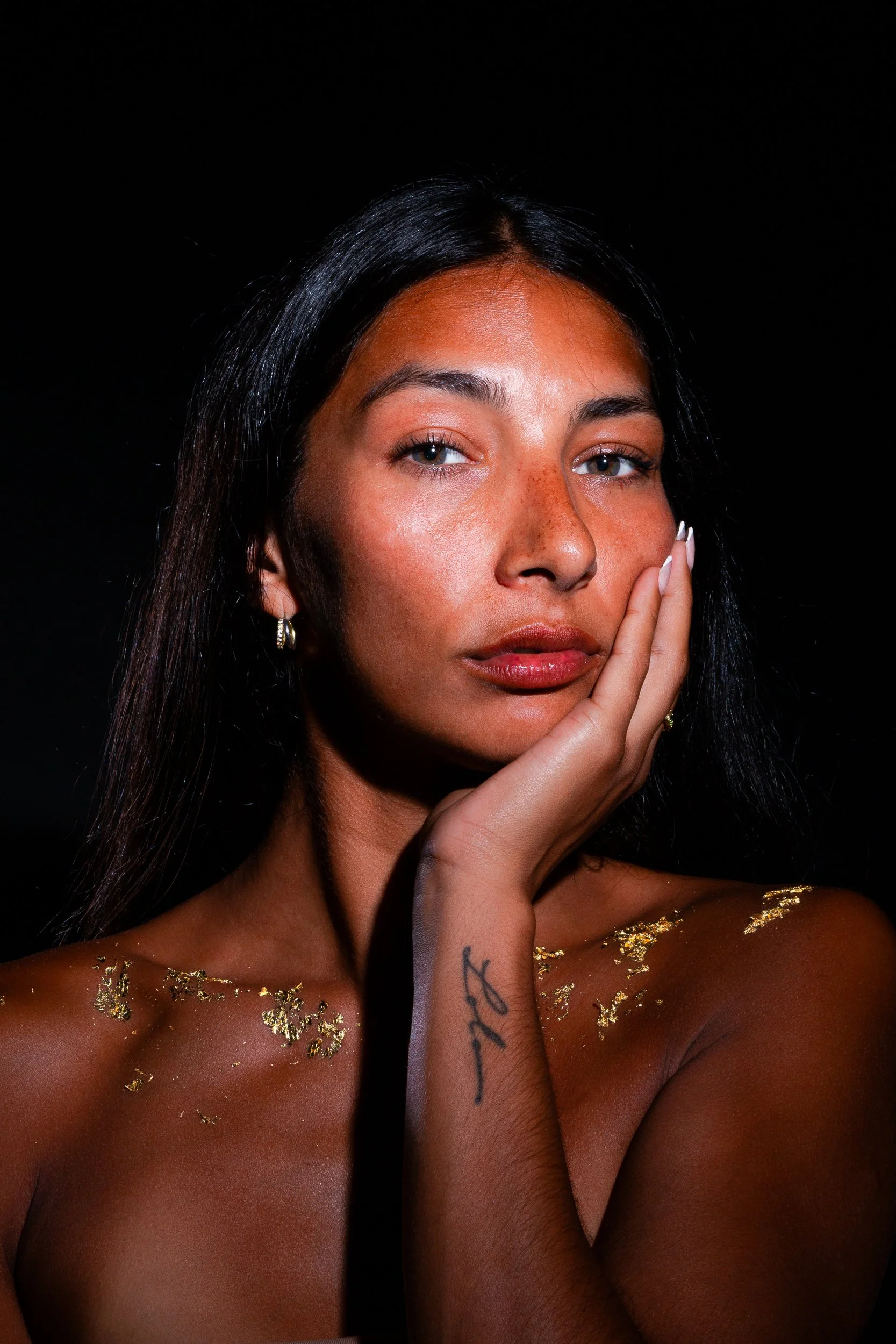A young woman with long dark hair, blue eyes, and freckles, touching her face gently. She has gold leaf on her shoulders and chest, and is wearing hoop earrings. The background is black.