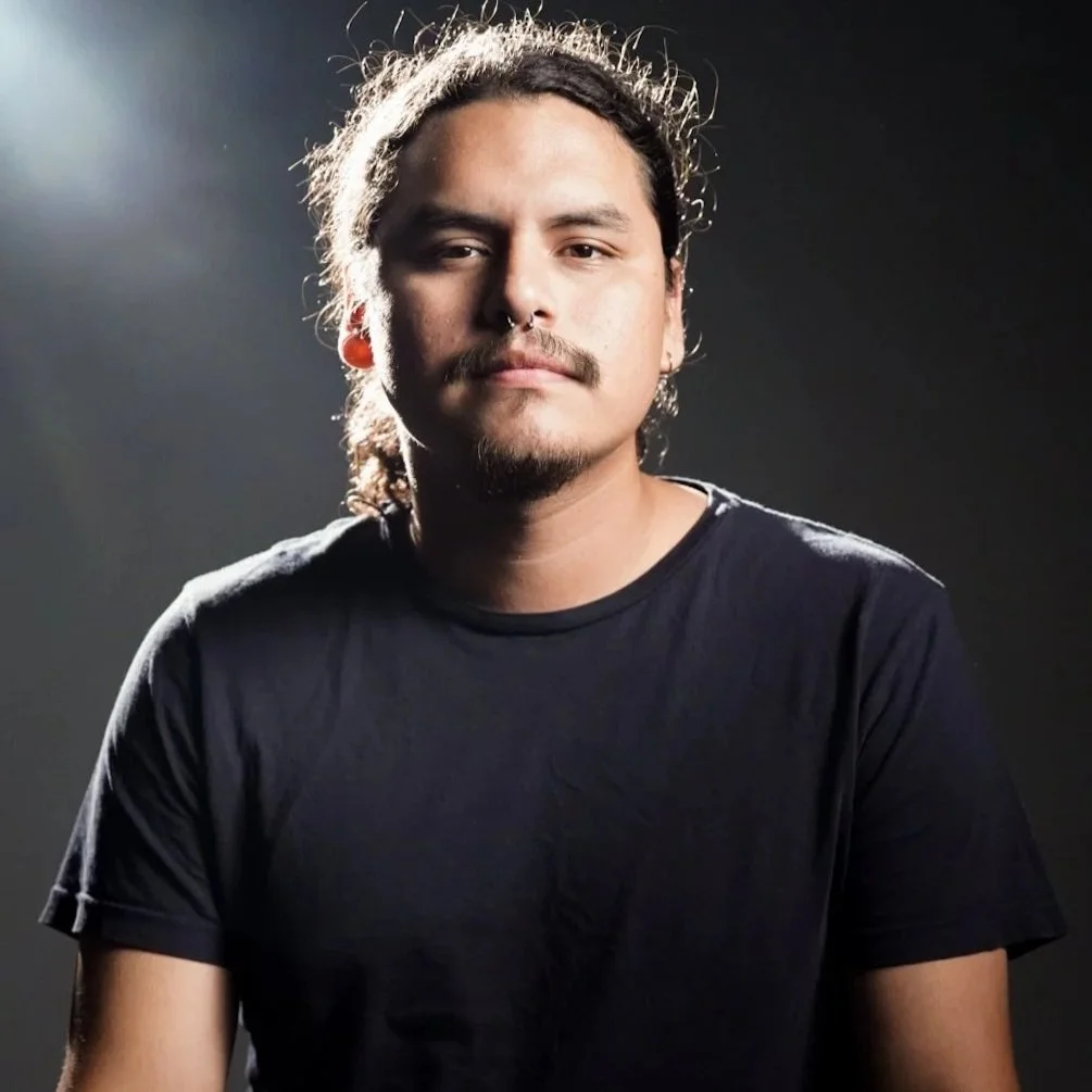 A young man with long hair, mustache, and goatee wearing a black t-shirt, posing against a dark background with a light source to his left.