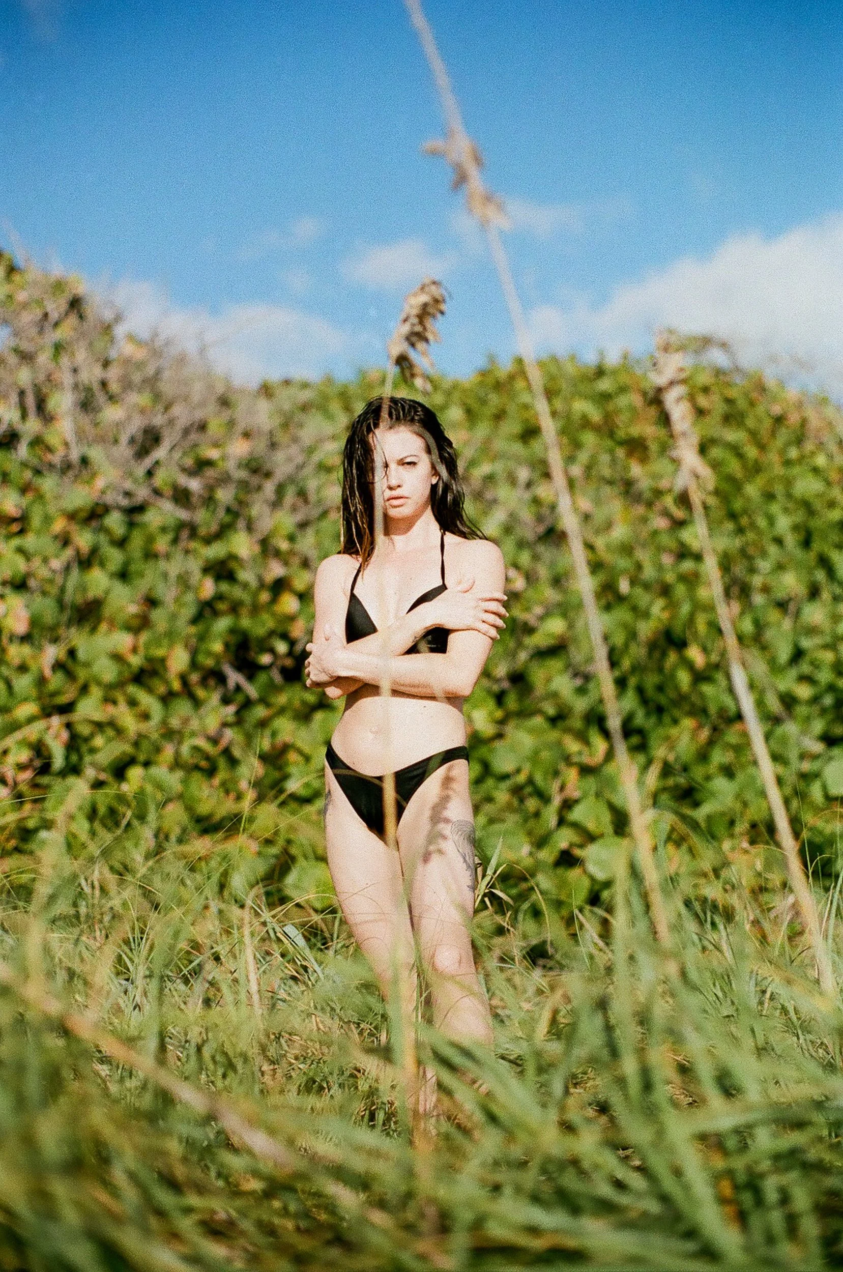 A woman with wet hair standing in a field of tall green grass and plants, wearing a black bikini, with a serious facial expression. There is a blue sky with some clouds in the background.