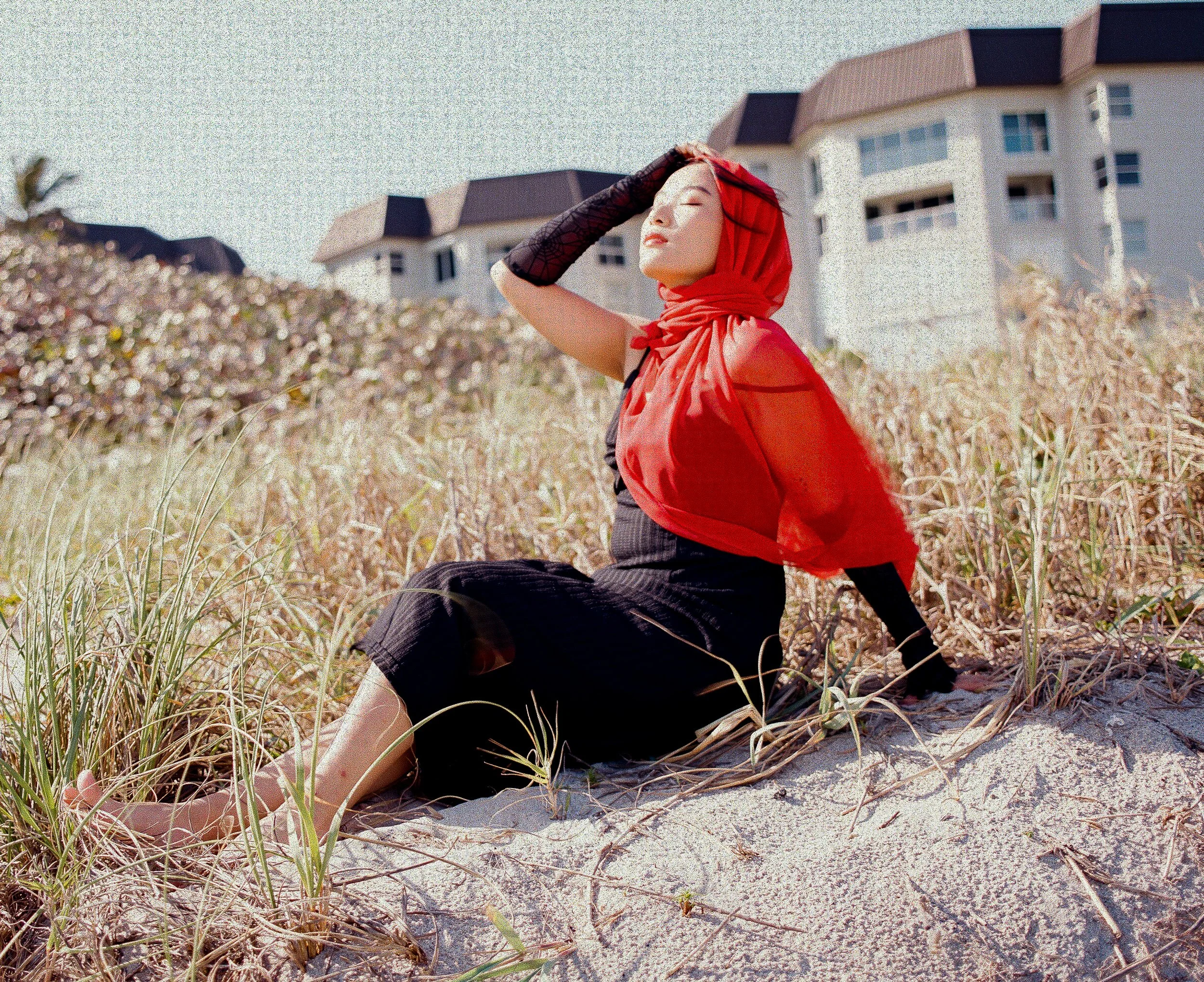 Woman in a red dress and black gloves sitting on sand dune with buildings in the background, eyes closed, her hand on her forehead, head tilted back.