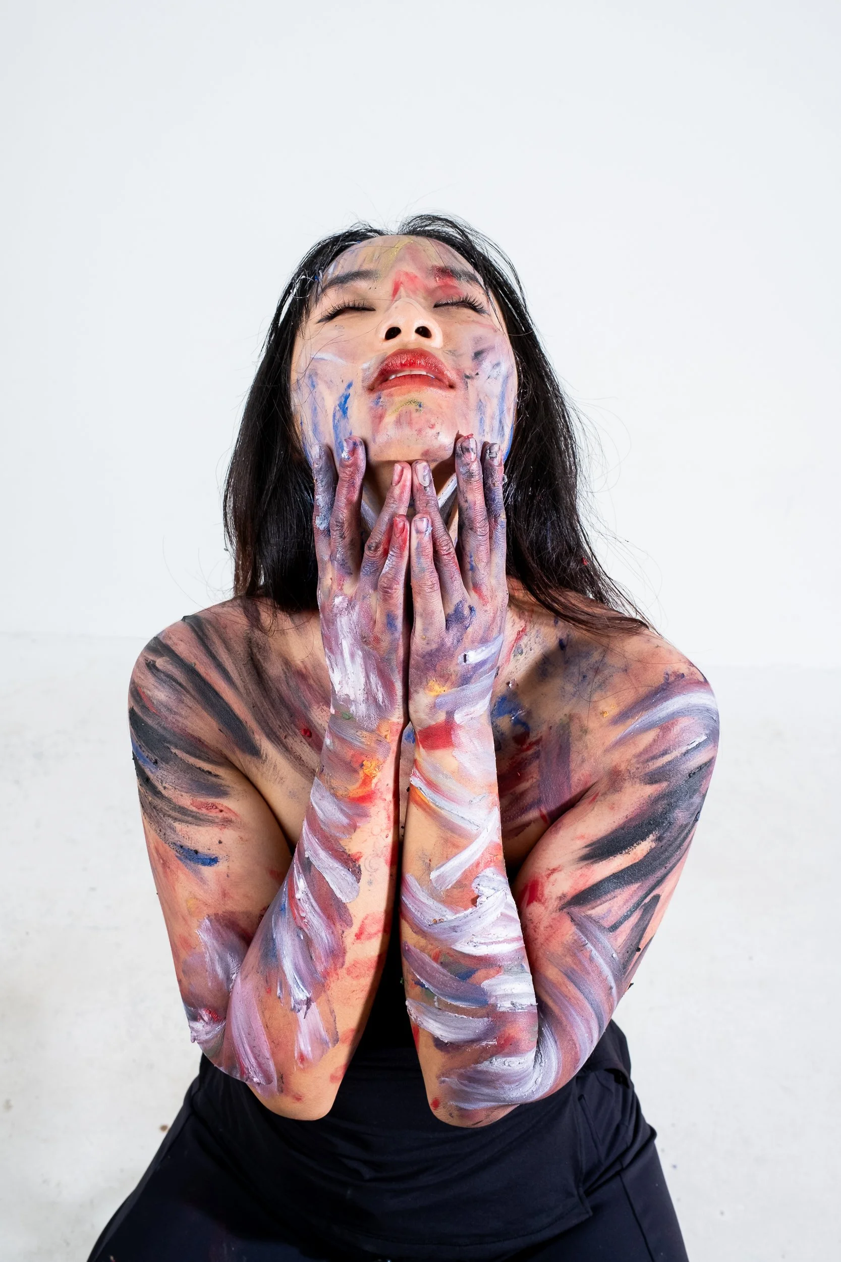Person with black hair covered in colorful paint, with paint on face, hands, arms, and shoulders, eyes closed, in front of a plain white background.