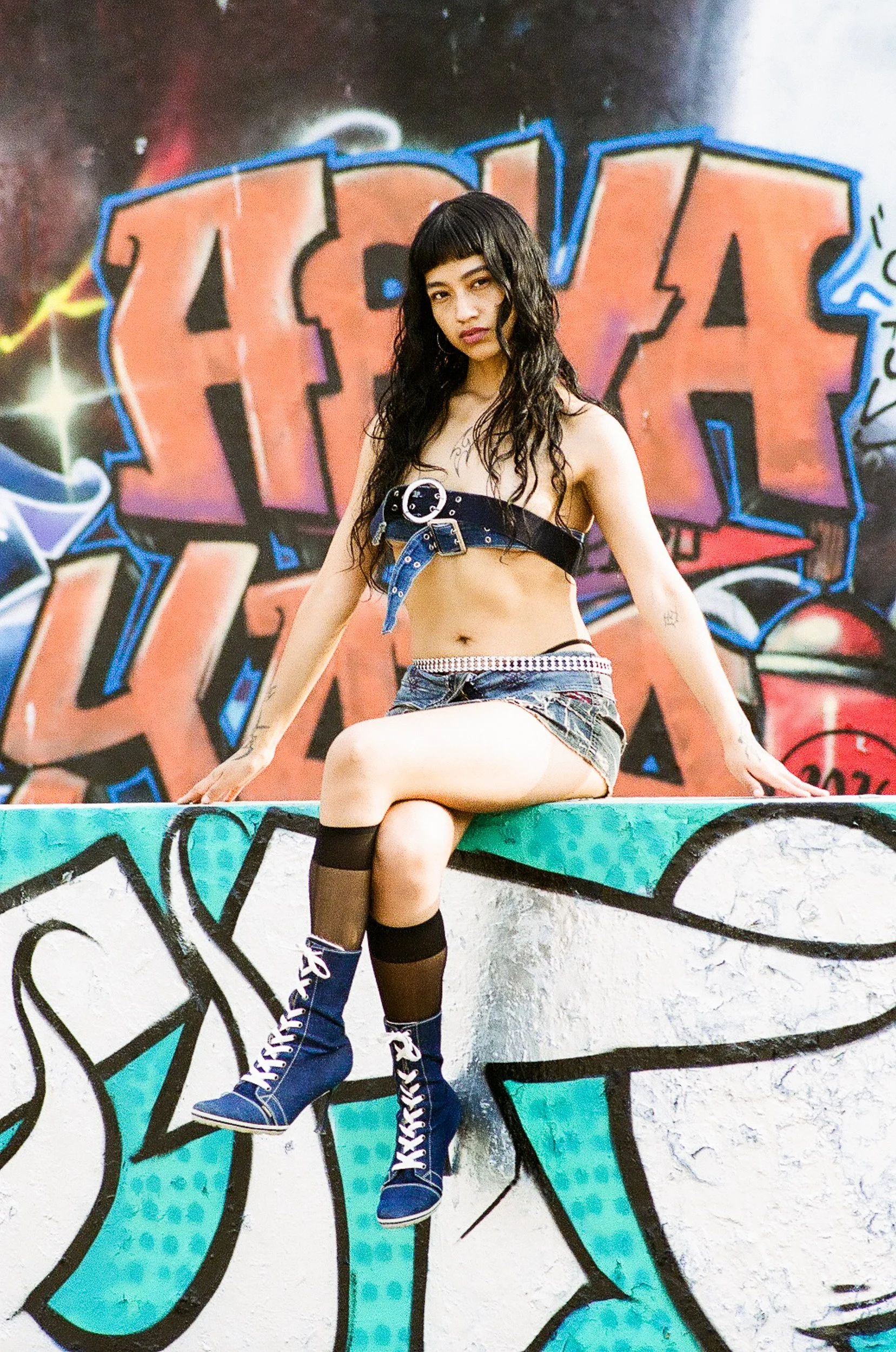 A woman with long dark curly hair, wearing a black strapless top with silver buckles, denim shorts, black thigh-high stockings, and blue lace-up boots, is sitting on a graffiti-covered ledge in front of a colorful graffiti wall.