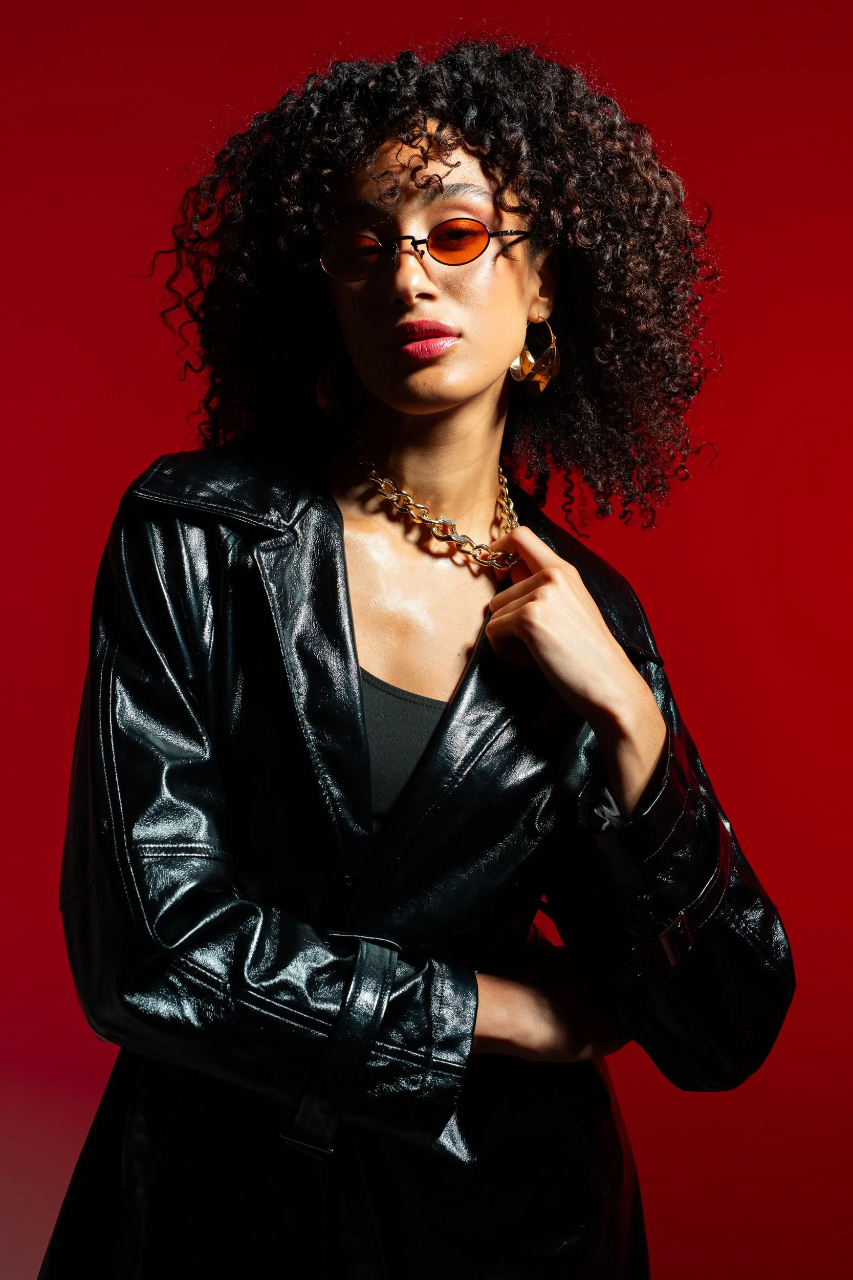 A woman with curly hair wearing orange sunglasses, gold earrings, a chunky gold chain necklace, a black top, and a black shiny leather jacket against a red background.
