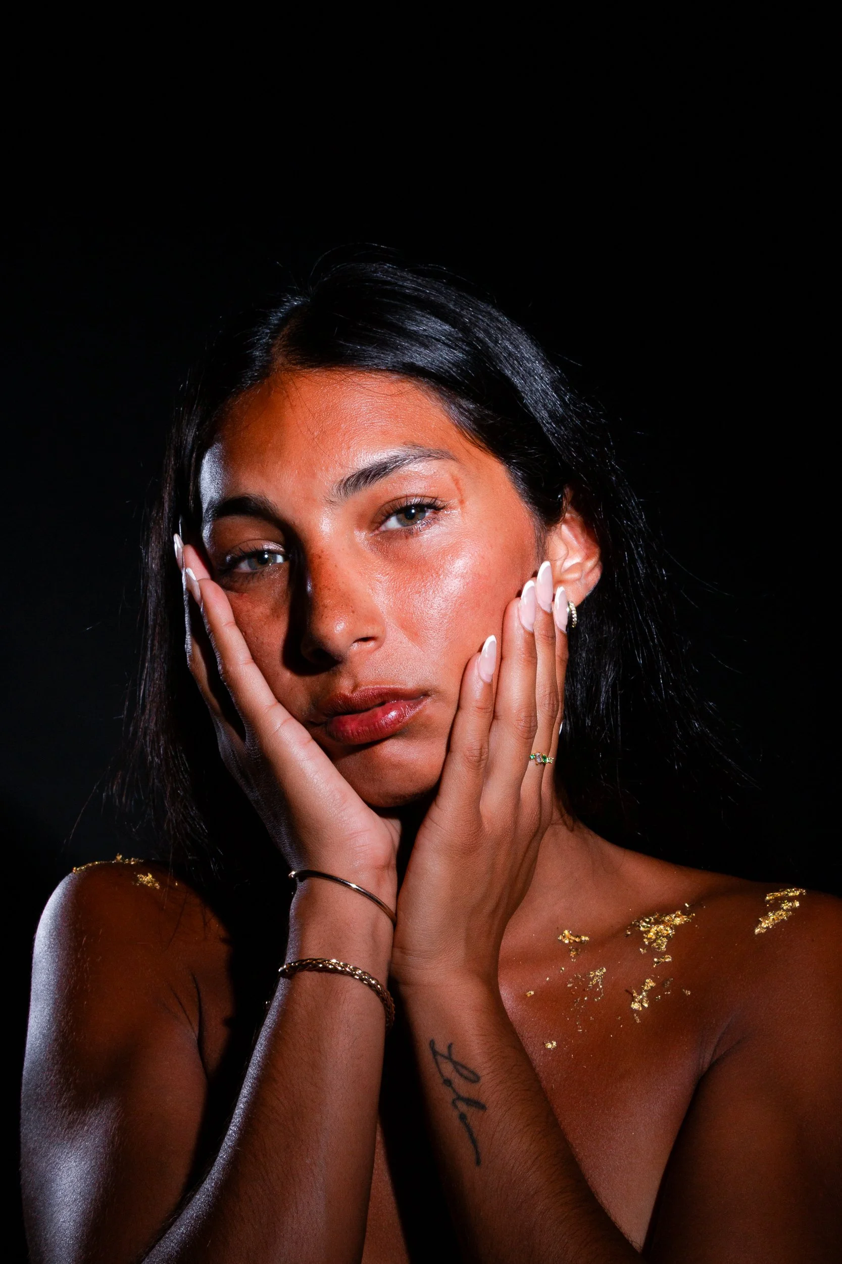 A woman with long black hair, tan skin, and blue eyes poses with her hands touching her face against a black background. She has some tattoos and is adorned with gold miniatures and jewelry with gold accents.