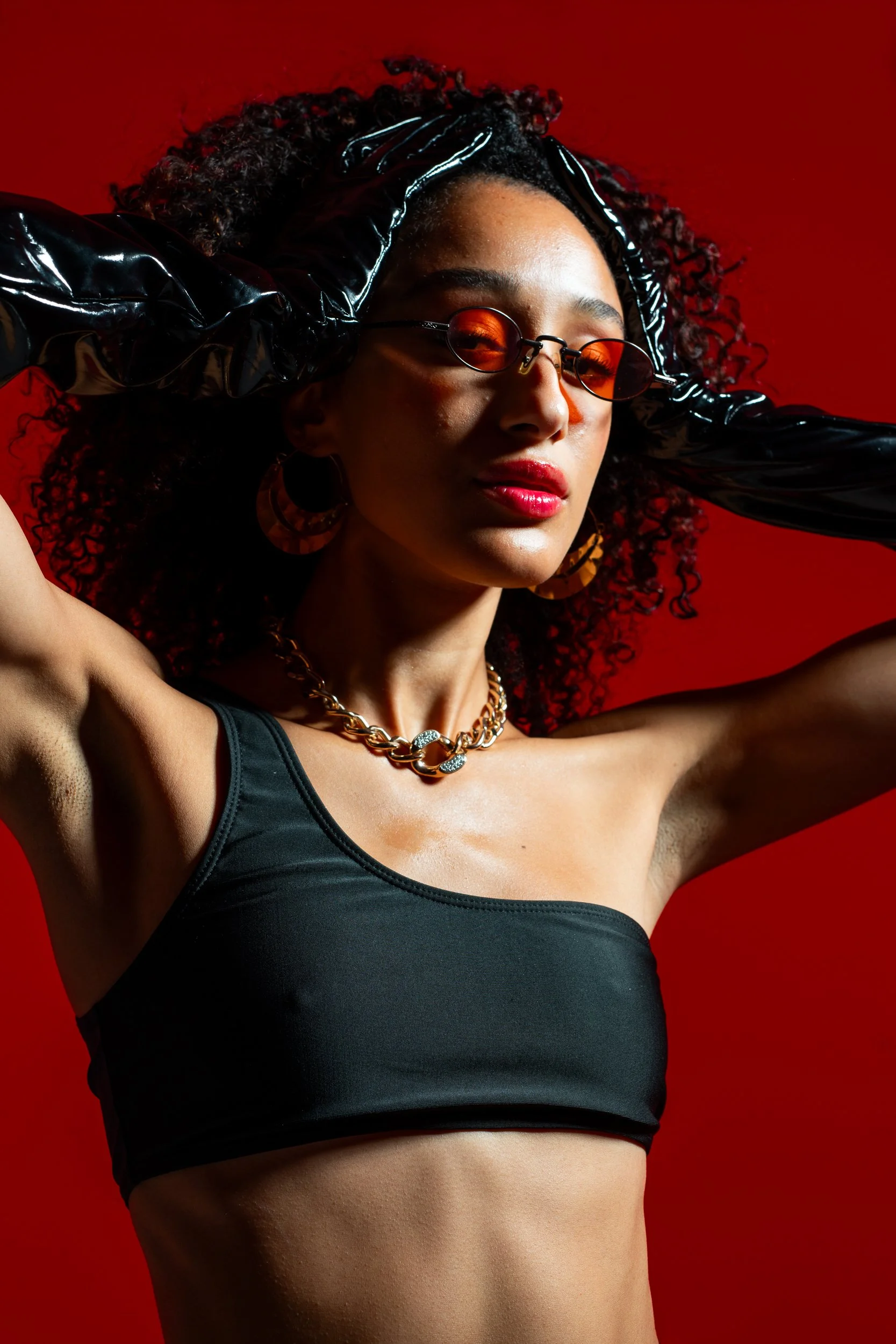 A woman with curly hair wearing orange-tinted sunglasses, large hoop earrings, a chunky gold chain necklace, a black tank top, black shiny gloves, and posing against a red background.