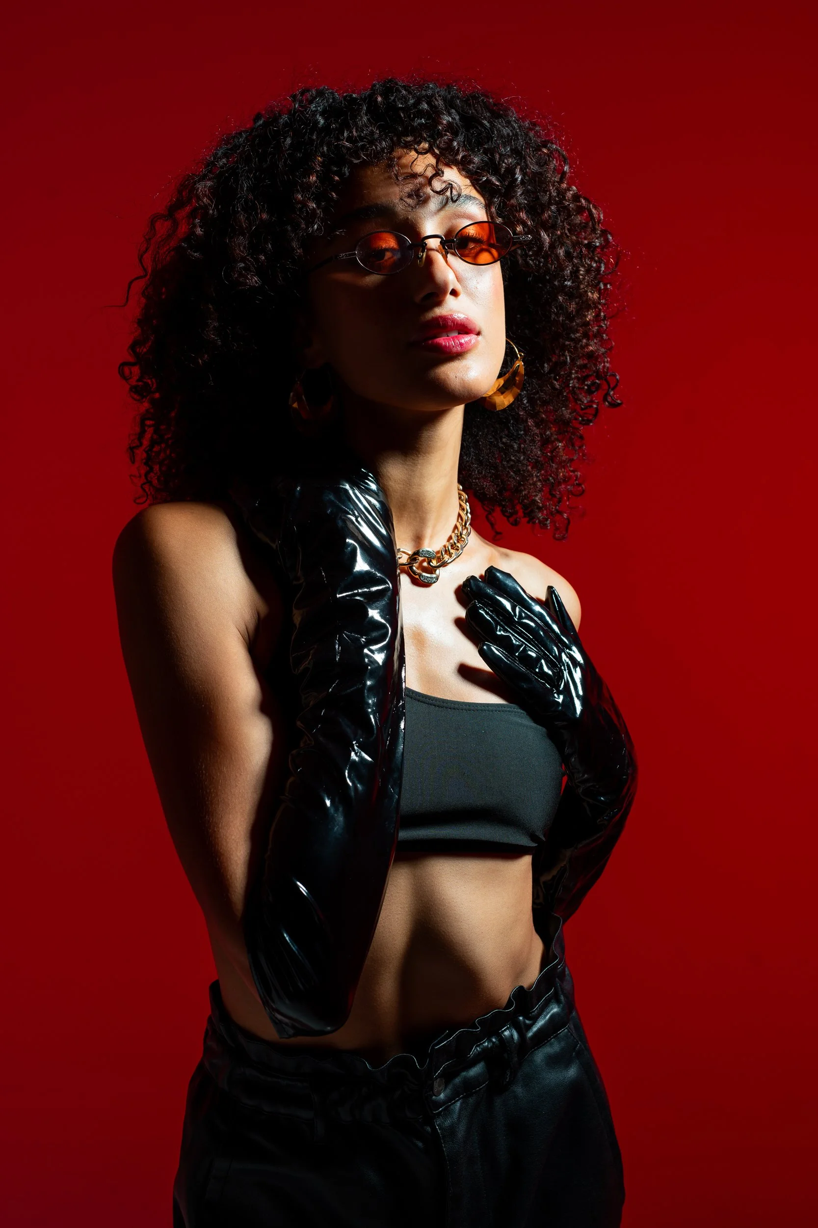 A woman with curly hair wearing orange sunglasses, gold jewelry, black gloves, a black crop top, and black satin pants poses against a red background.