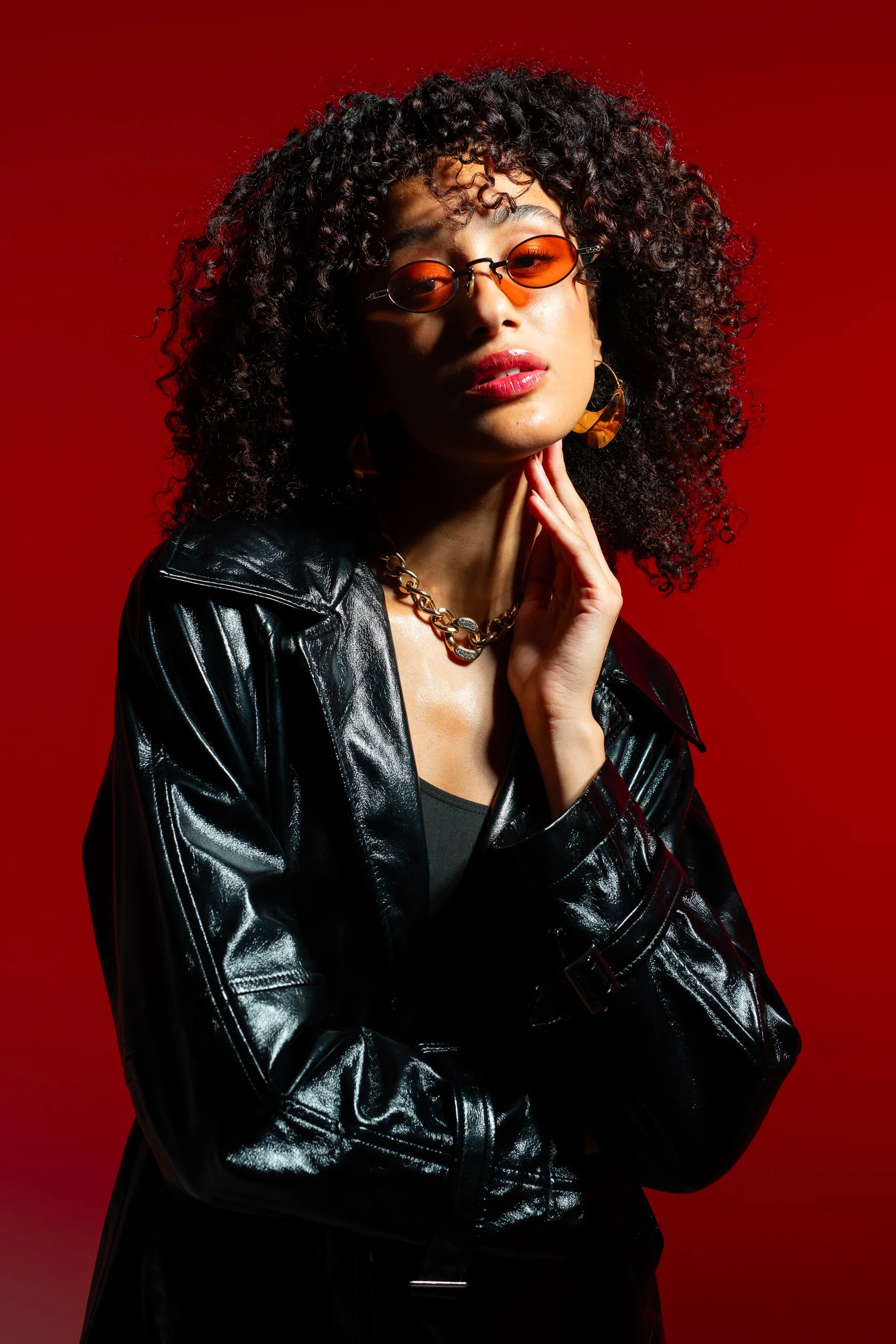 A woman with curly black hair wearing orange tinted sunglasses, a gold chain necklace, large gold earrings, and a black leather jacket against a red background.