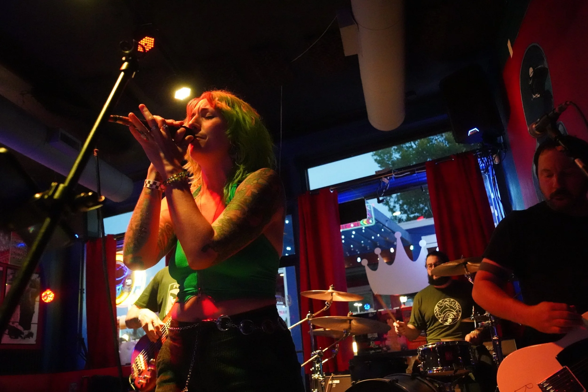 A woman with green and red hair singing into a microphone in a dimly lit bar or club, with a drummer and a man playing guitar in the background, illuminated by colorful stage lights.