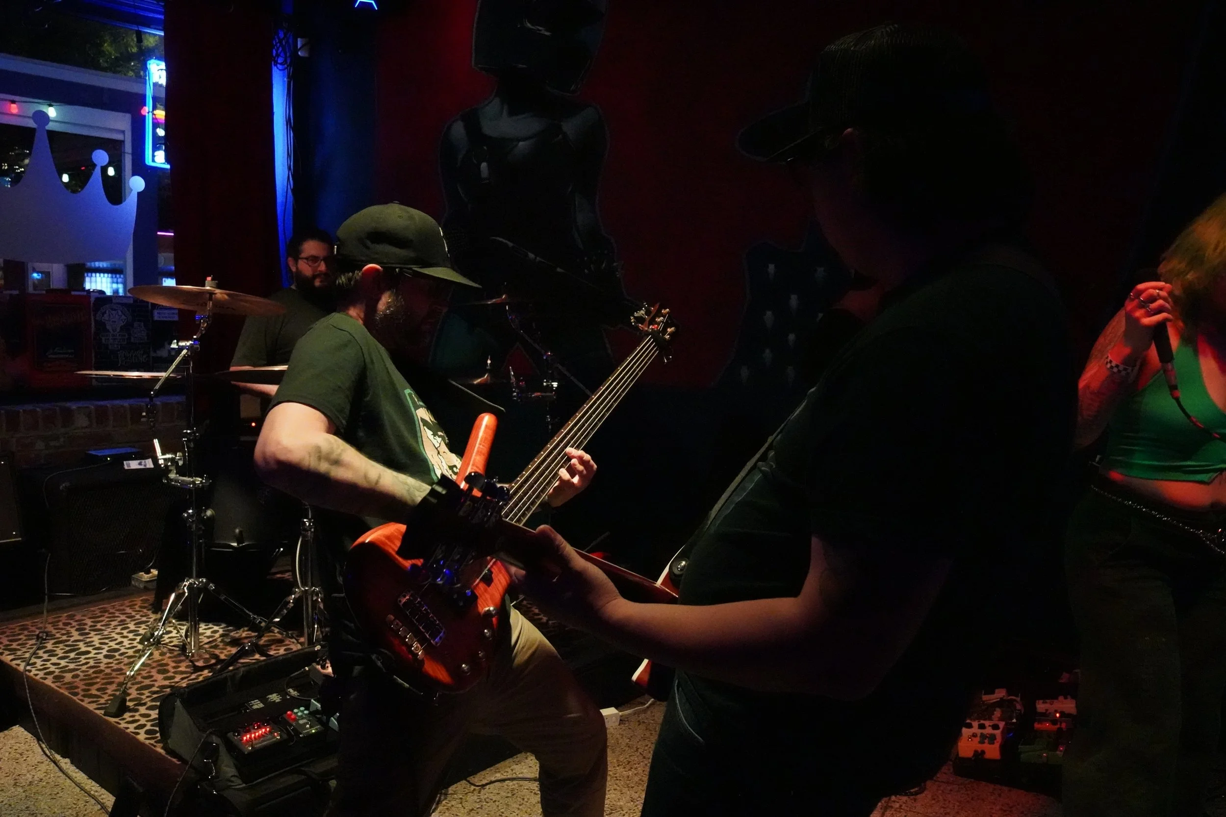 A dimly lit indoor music rehearsal or performance space with a female singer holding a microphone, a man playing an electric bass guitar, a drummer, and a person with headphones. Neon and colored lights are visible in the background.