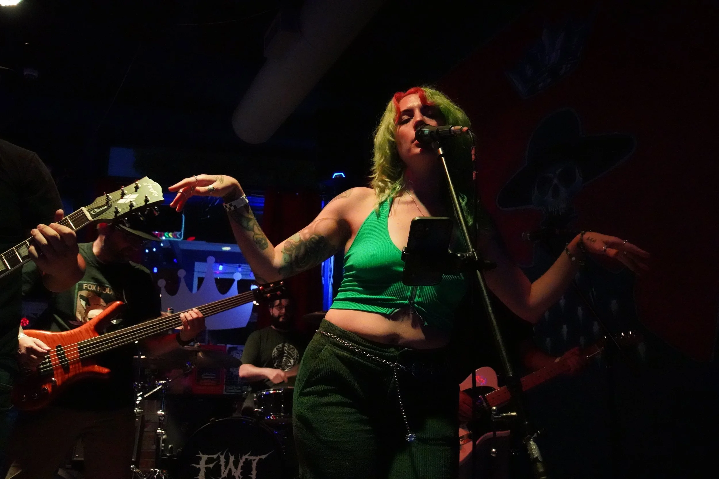 A woman with green hair dyed with red streaks singing into a microphone on stage while playing guitar. She is wearing a green crop top and black pants with chains. Several musicians and instruments are visible in the background.