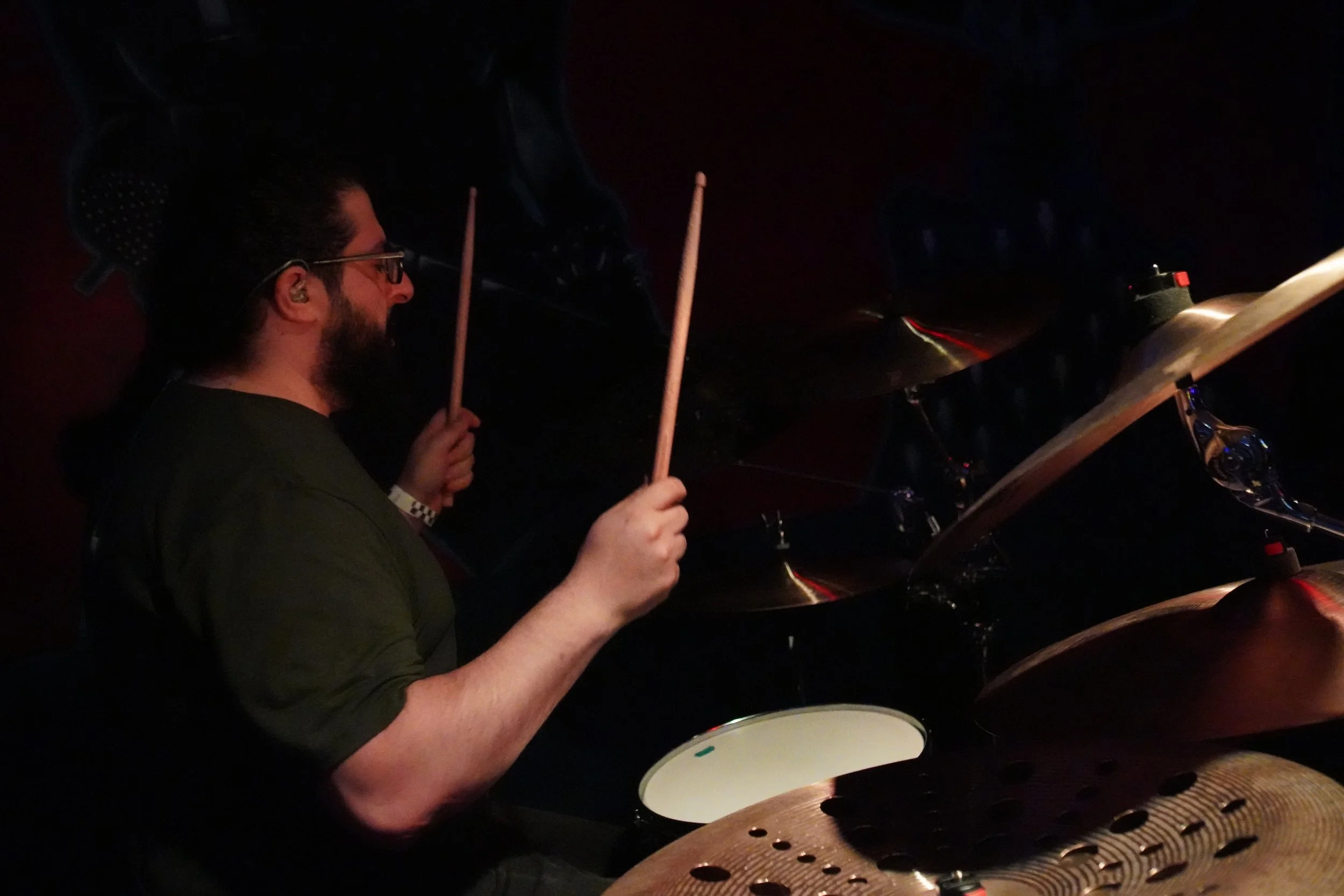 A man playing drums in a dimly lit room, seen in profile, holding drumsticks, with a sheet of music and drum hardware nearby.