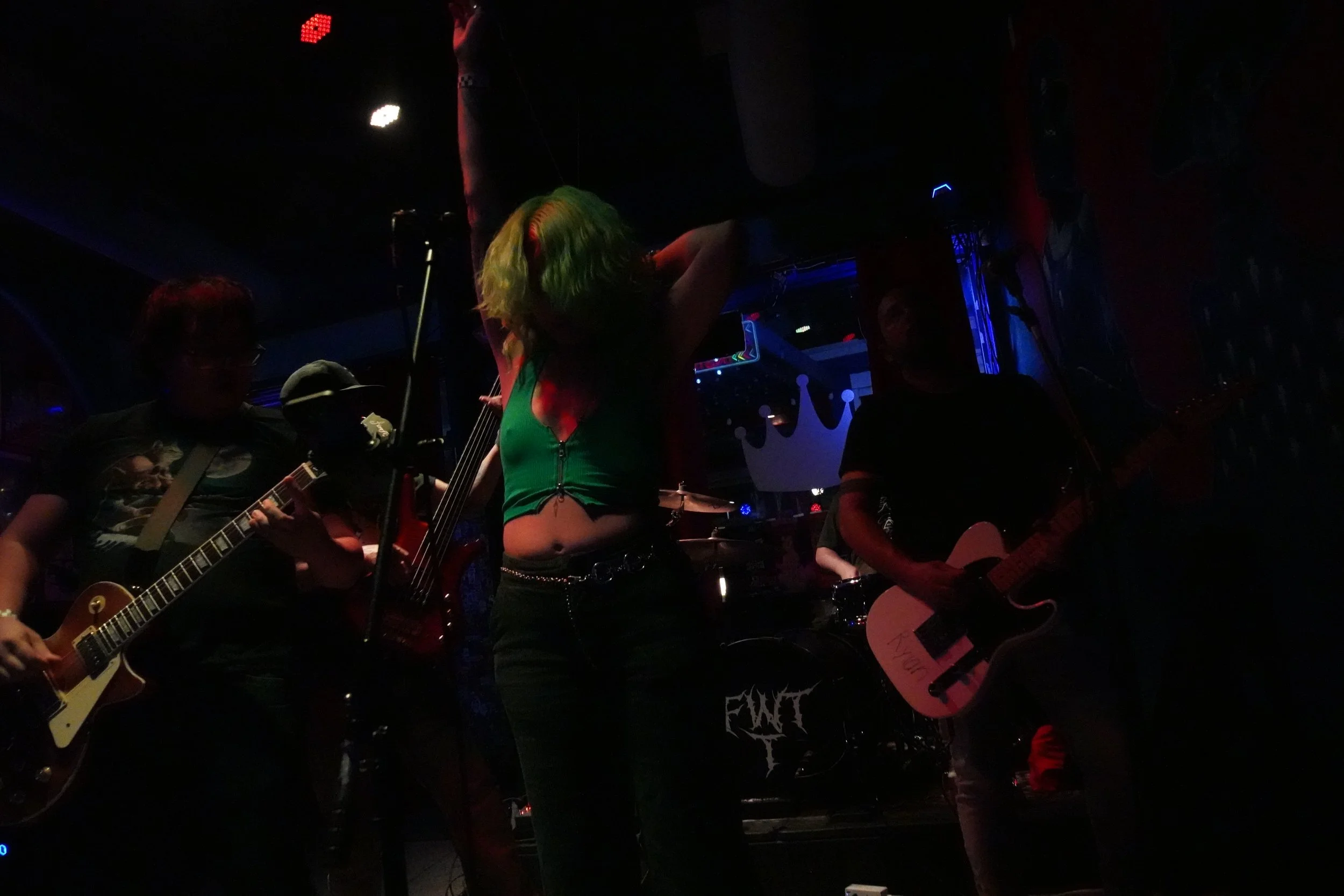 A dimly lit stage with band members performing, including a woman with bright green hair and a green top, playing guitar, and other musicians playing guitars and drums, with colorful lights and a dark background.
