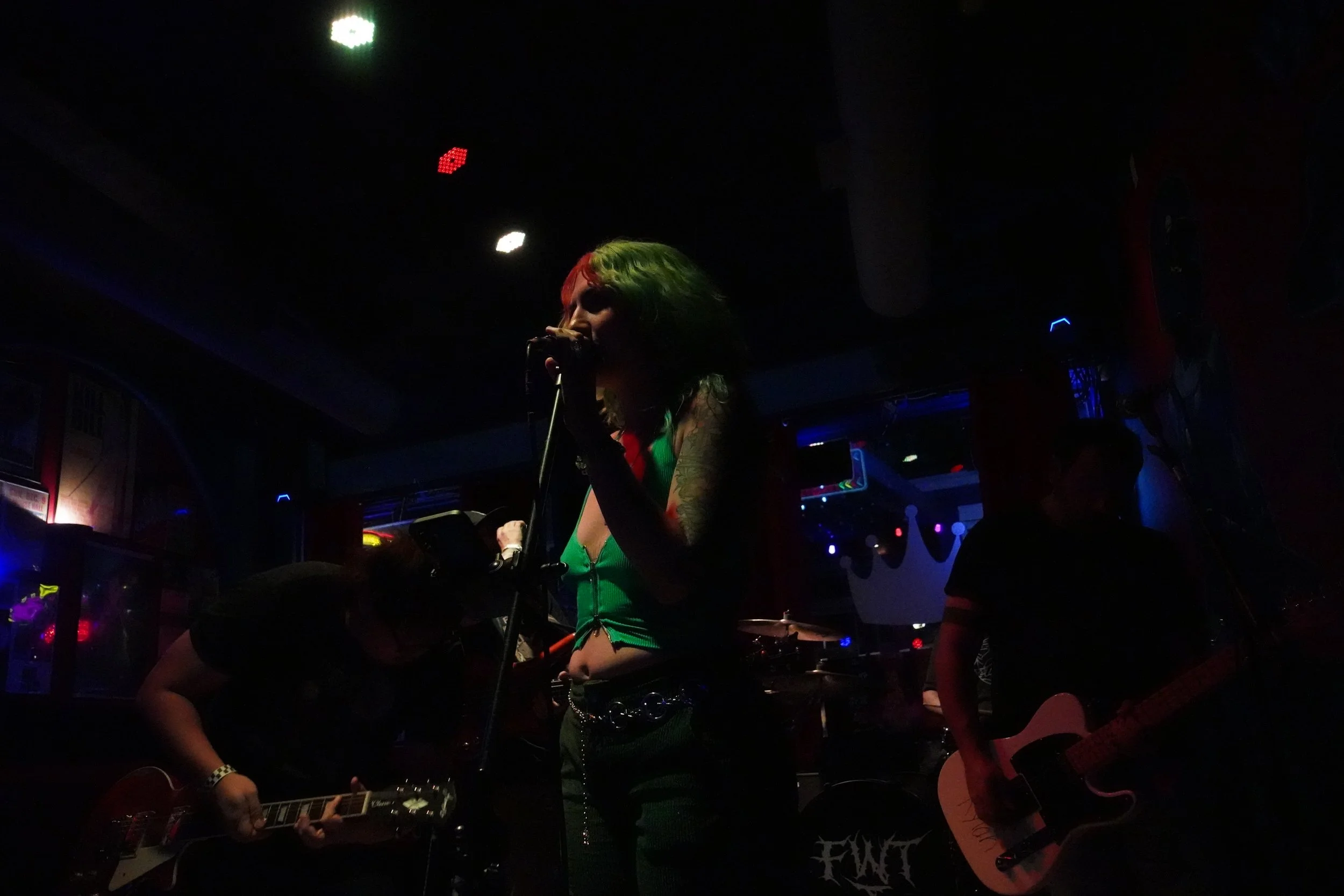 A dark scene with a woman singing into a microphone, with colorful hair and tattoos, accompanied by people playing guitars, in a dimly lit venue with neon and stage lights.