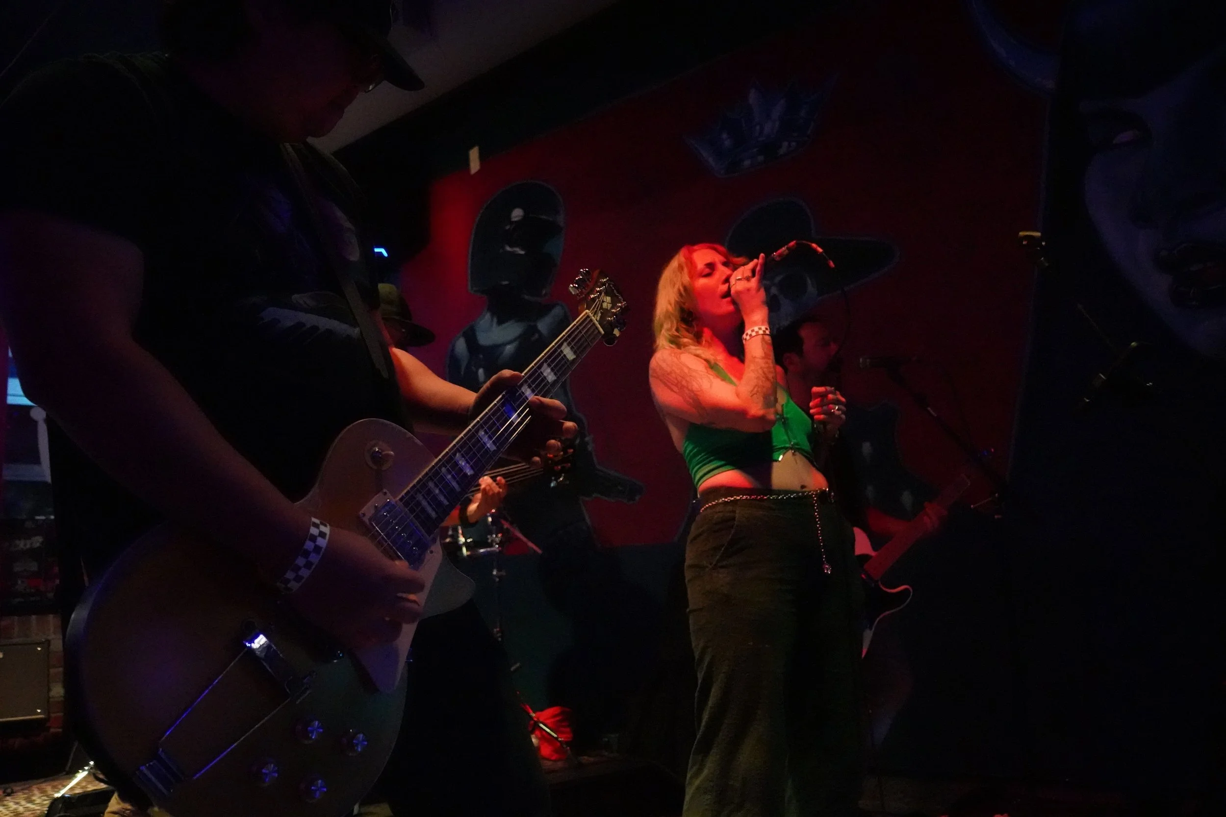 A woman singing into a microphone and a man playing guitar on stage, with other band members partially visible, in a dimly lit live music venue.
