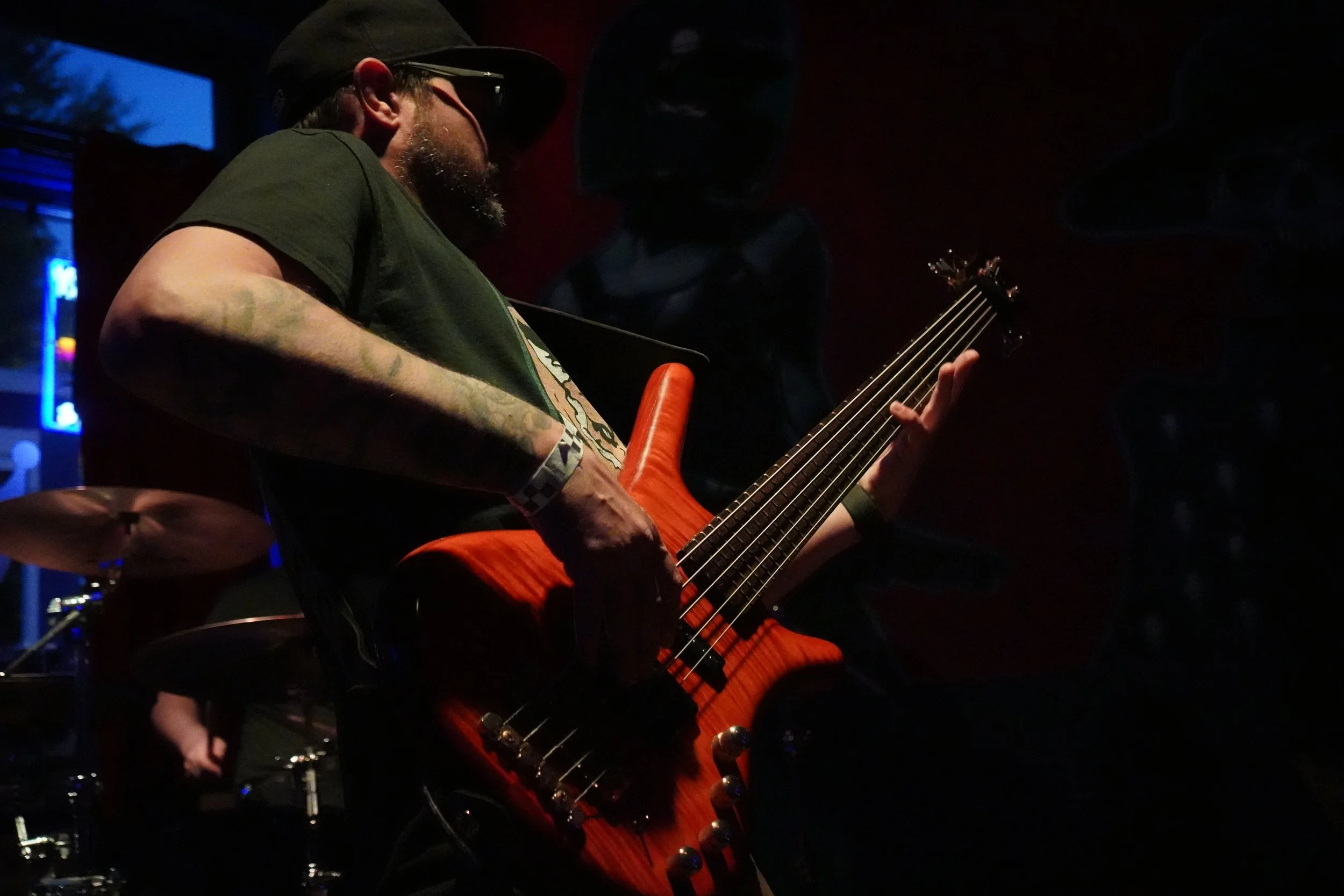 A man with tattoos and a beard playing an orange electric bass guitar in a dimly lit venue.