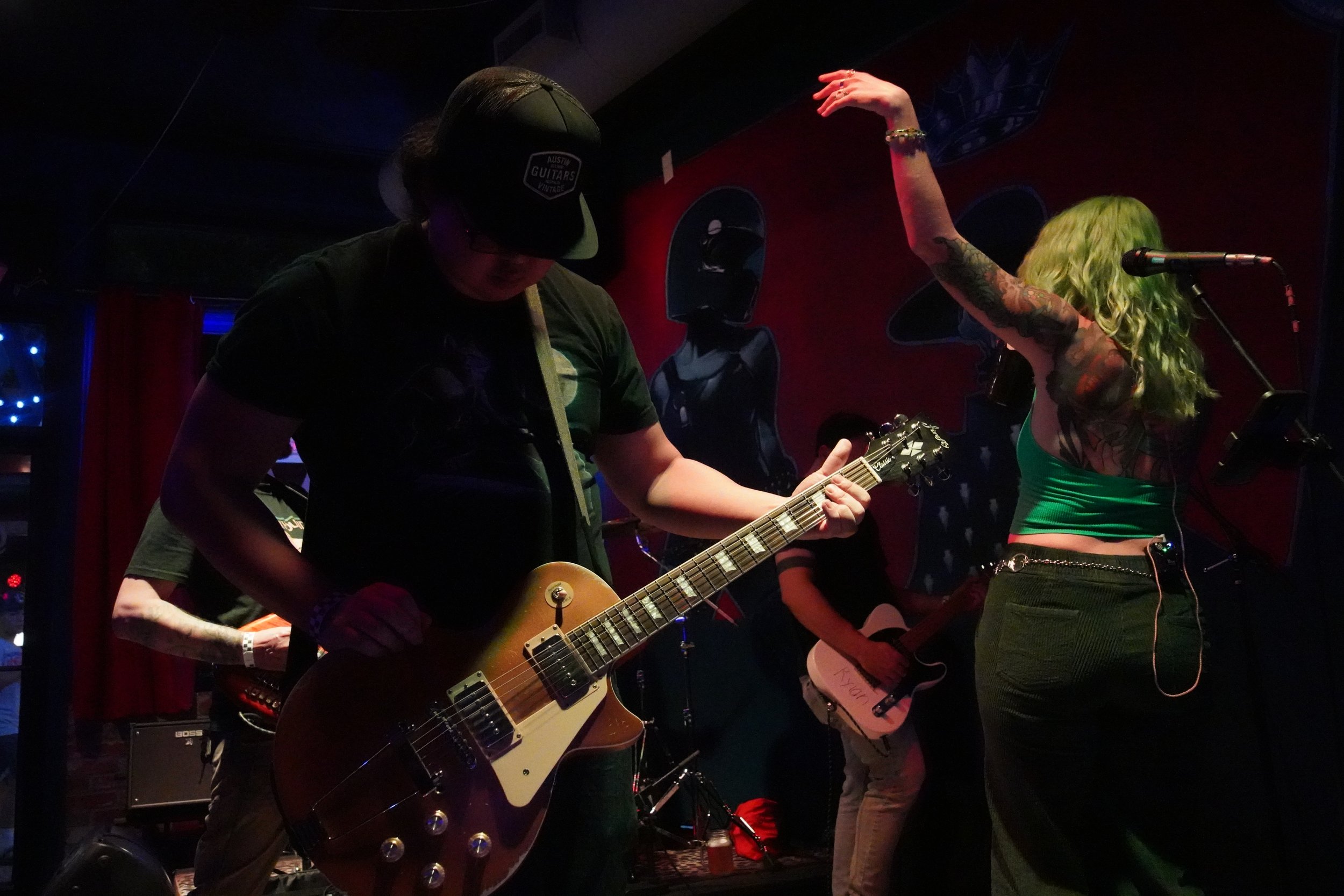 Musicians performing on stage with guitars, one woman with green hair and tattoos singing, and a man wearing a cap playing the guitar, in a dimly lit venue.