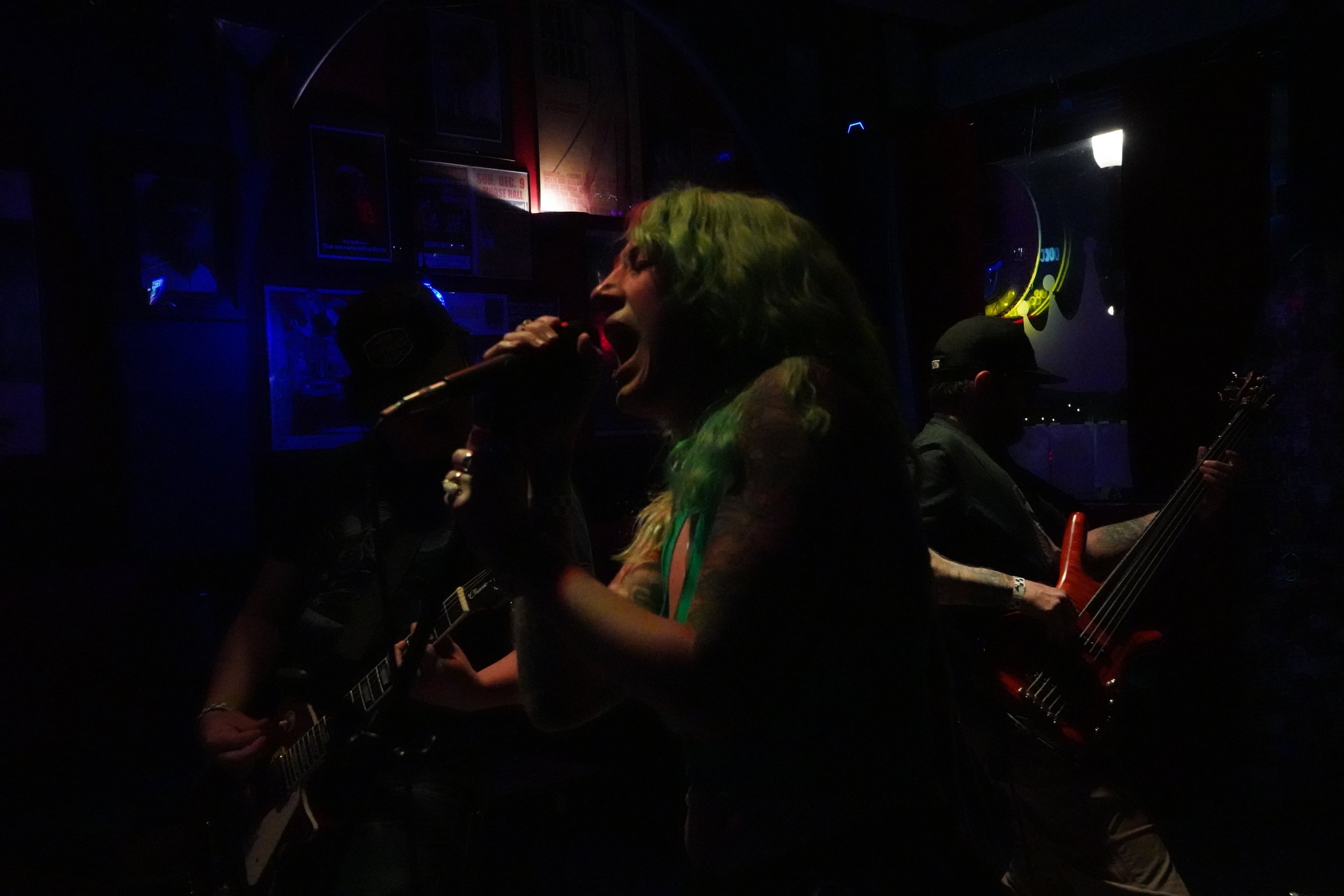Woman with green hair singing into a microphone in a dimly lit venue, accompanied by musicians playing guitars.