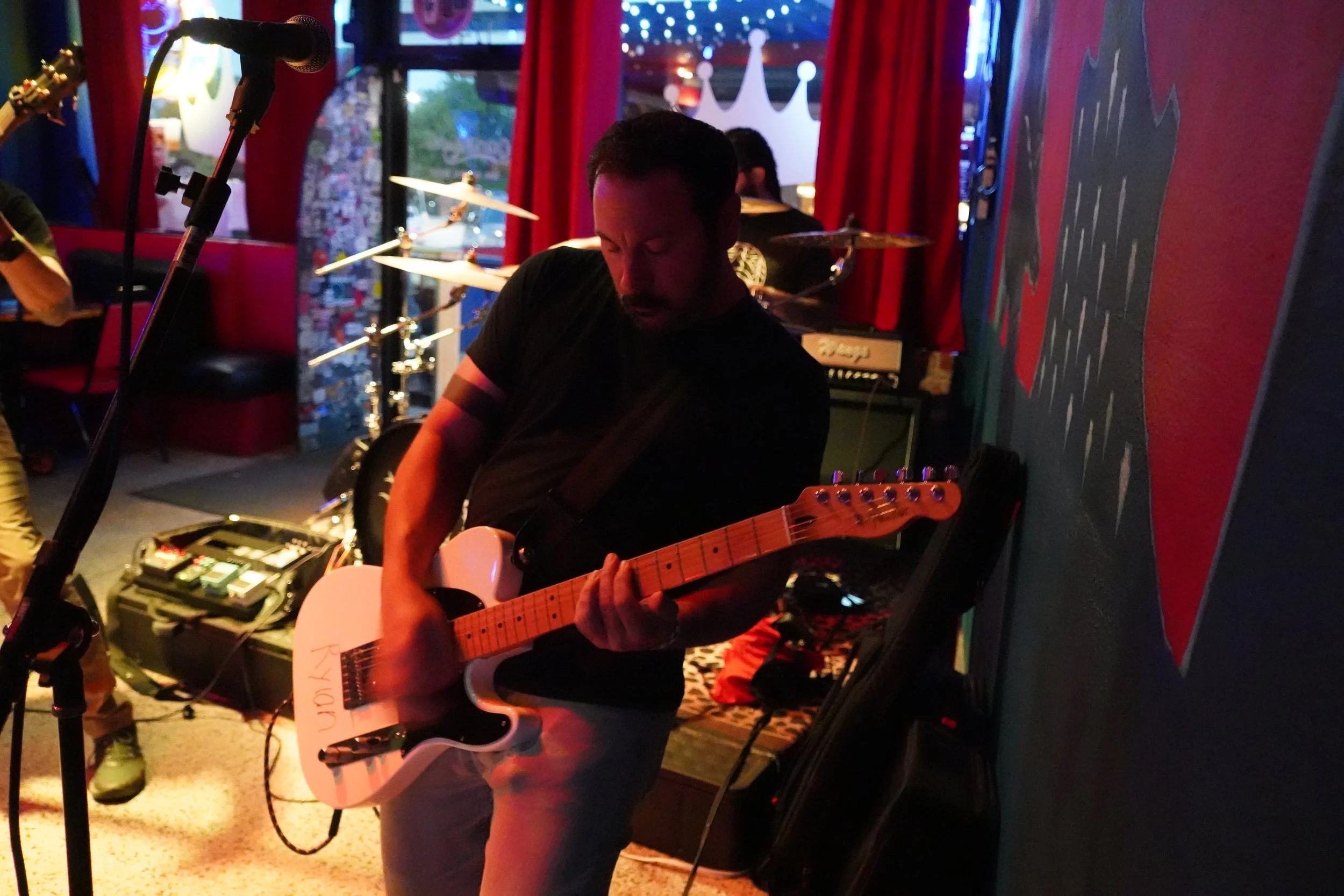 A man playing an electric guitar at a dimly lit venue with red curtains and musical equipment around him.