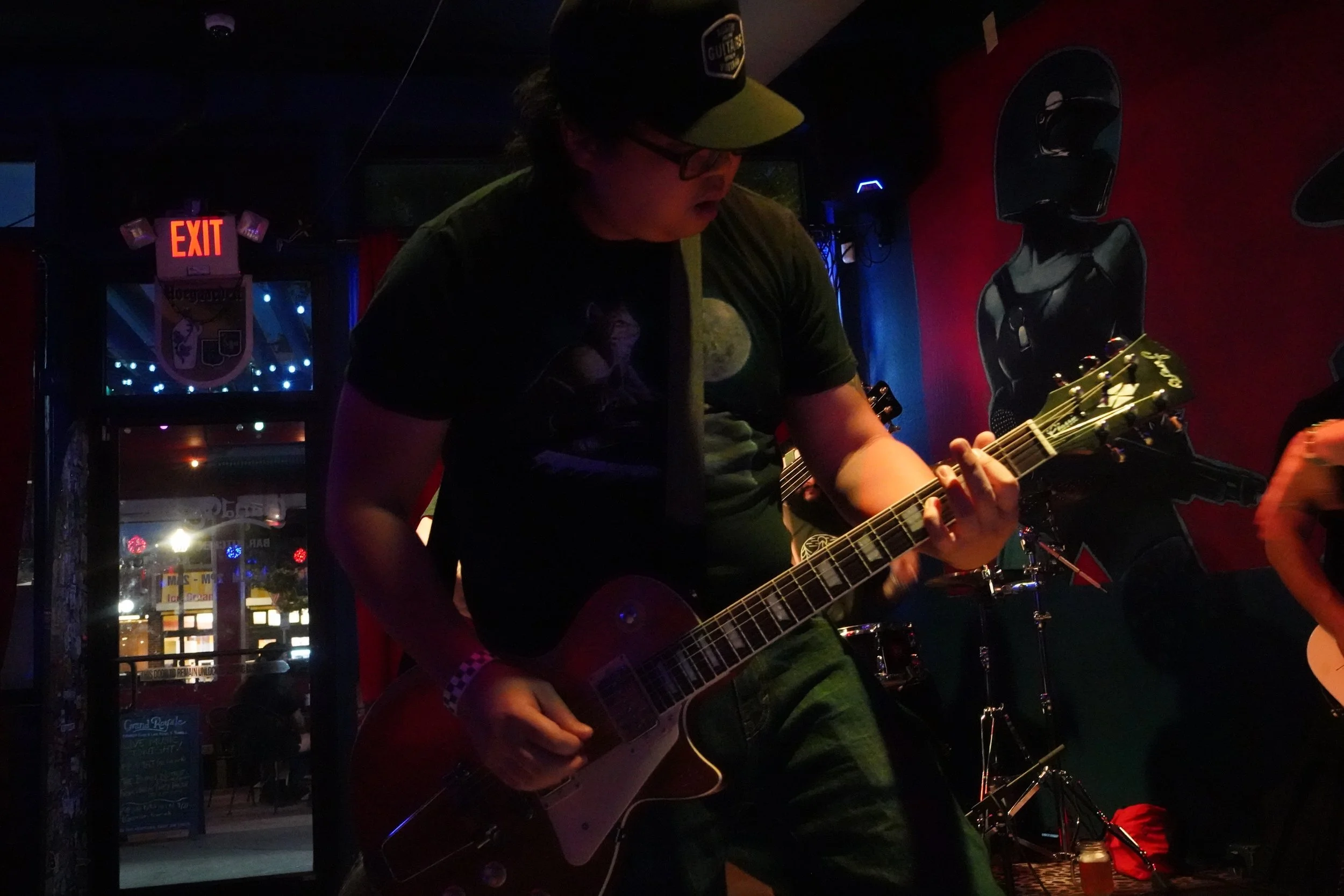 A person wearing glasses and a cap playing an electric guitar in a dimly lit venue with red and blue lighting.