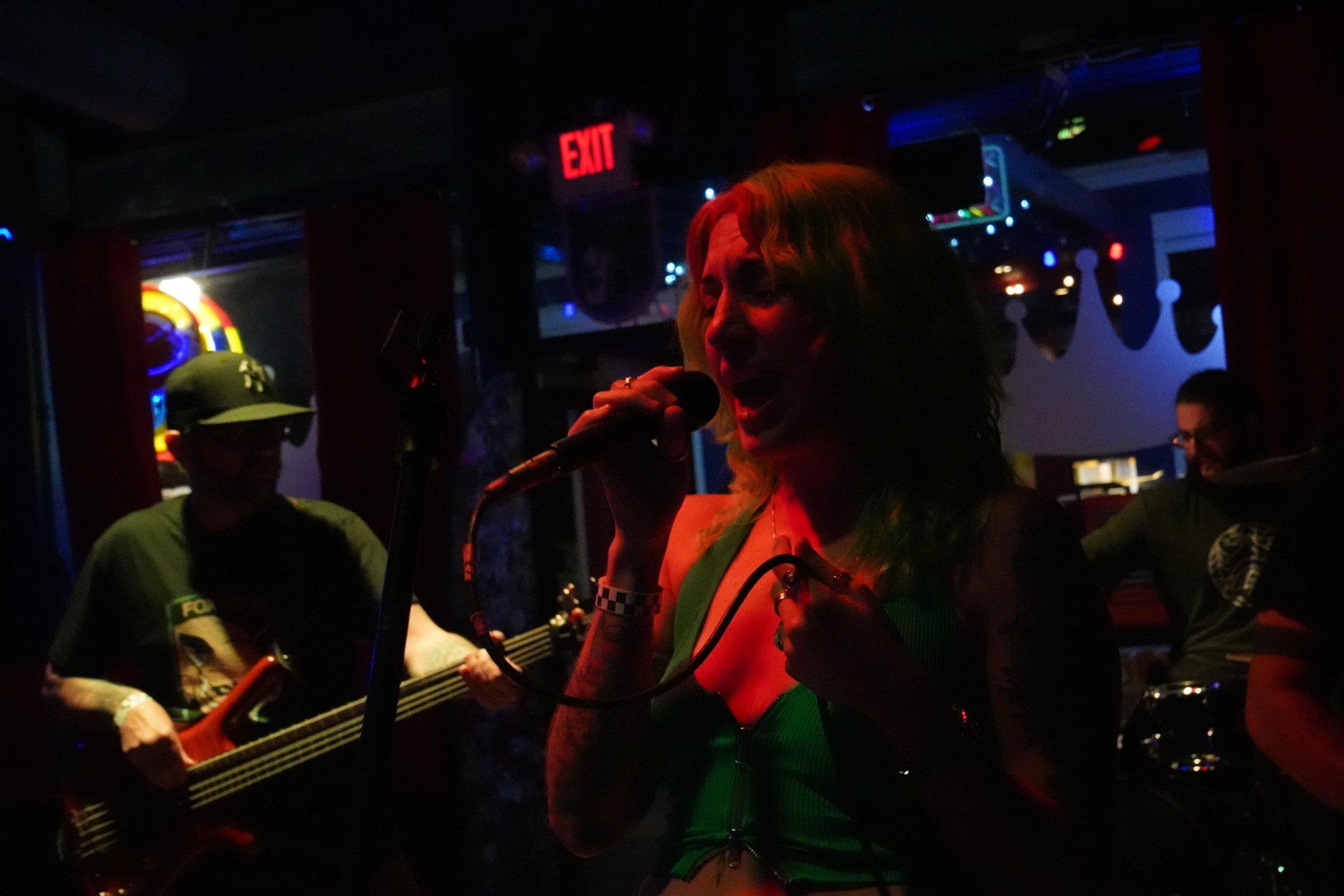 A woman singing into a microphone at a dimly lit venue, with a band playing guitar in the background and colorful lights.