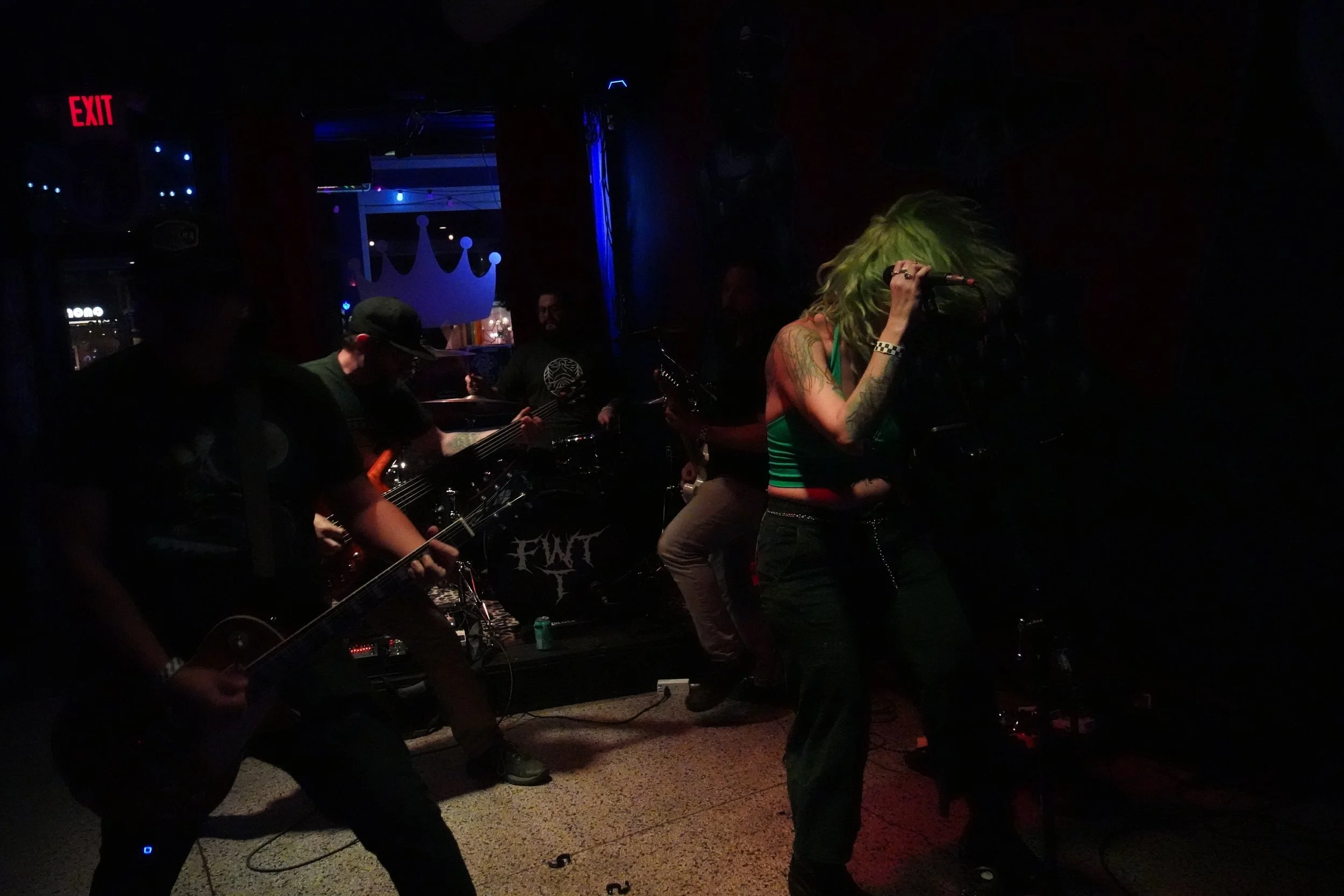 A musical performance in a dark venue with four musicians playing instruments and a woman singing into a microphone, with blue and red lighting and a crown decoration in the background.