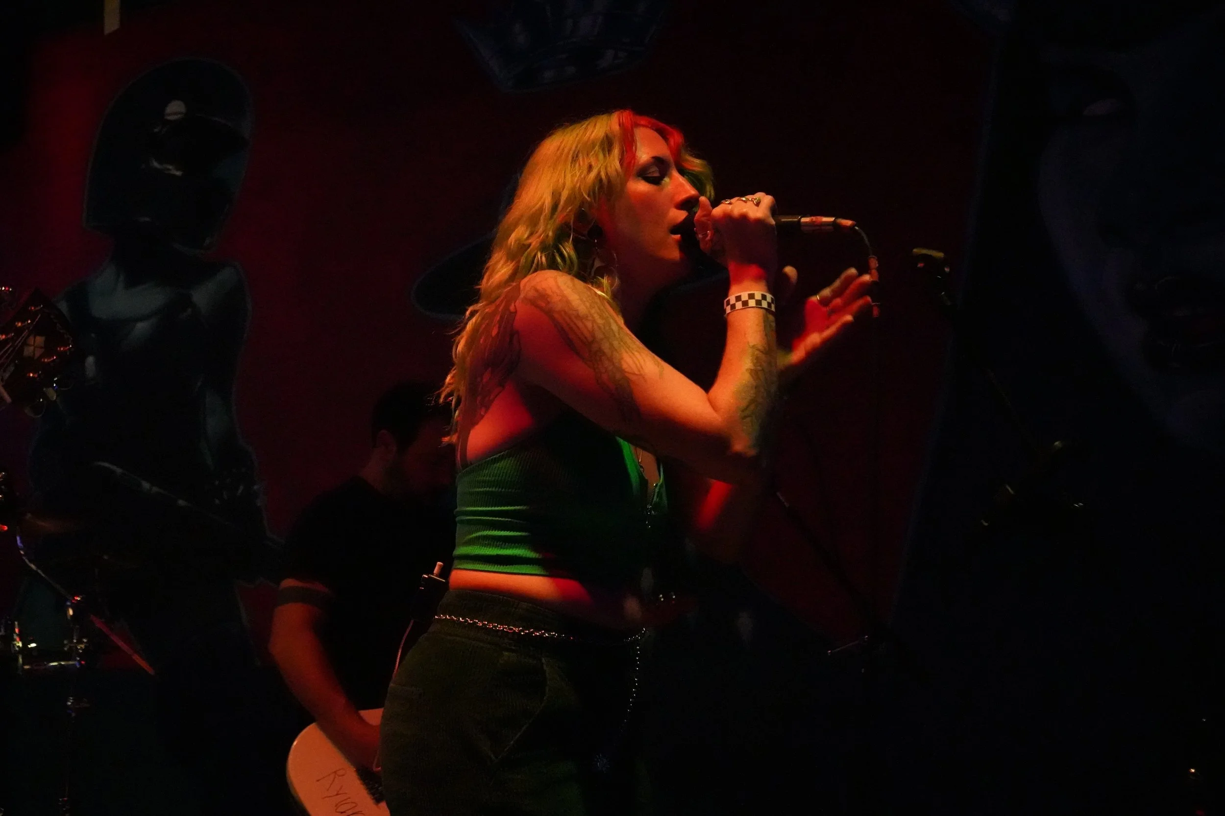 A female singer with long blonde hair and tattoos on her arms, wearing a black hat and a green top, singing into a microphone on stage with red and green lighting.