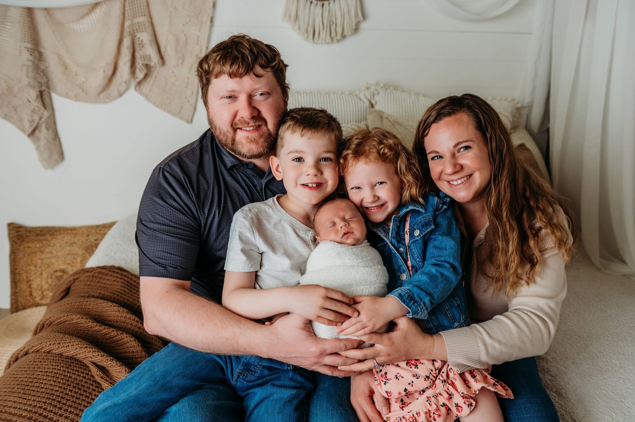 A family of six sitting on a couch indoors, smiling and embracing each other. The family includes two adults, three children, and a newborn. The background has a cozy and decorated setting.