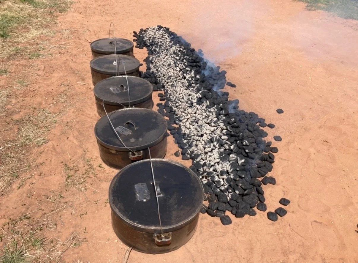 A series of five black heating or cooking containers with metal wires connecting them in a straight line, placed on dry ground next to a circular arrangement of green, white, and black rocks.