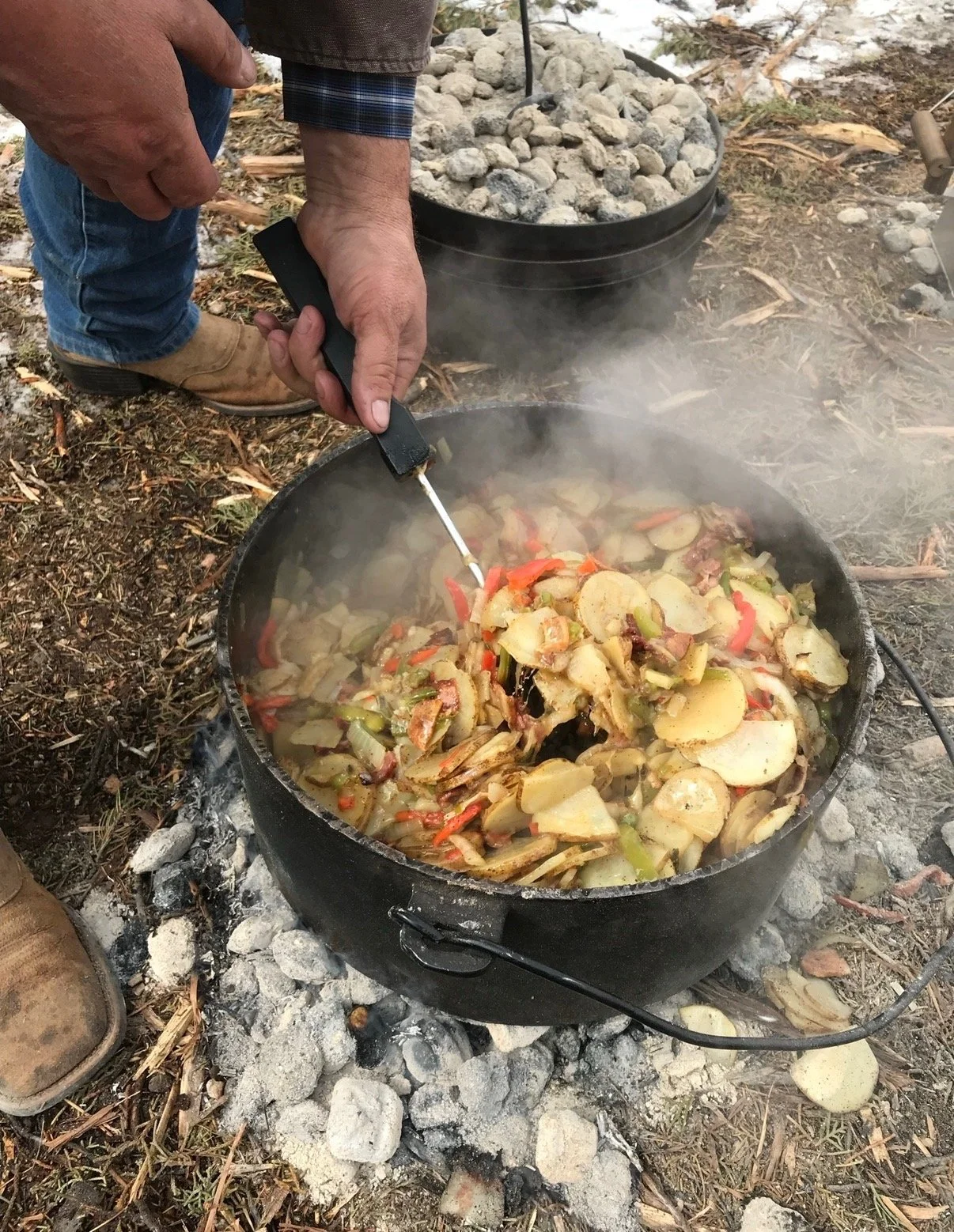 Someone stirring a large pot of cooked vegetables outdoors over a campfire, with another pot of stones or rocks in the background.