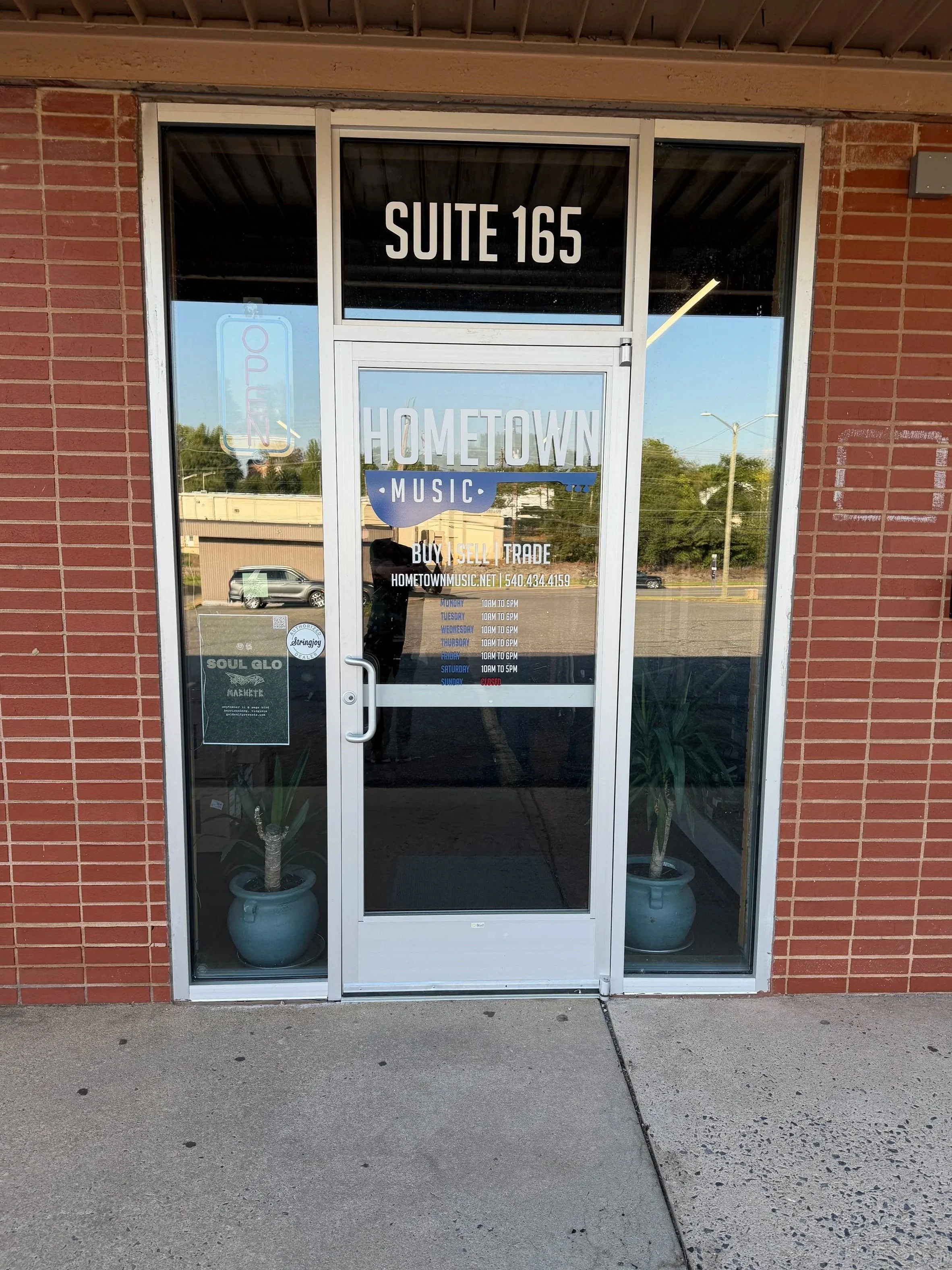 The image shows the entrance to a store called Hometown Music, with a brick wall on either side and glass doors. The store's sign indicates it's Suite 165. There are potted plants inside the store near the entrance. The reflection on the glass shows cars parked outside, a road, and some greenery with trees. The store's hours and contact information are also visible on the door.