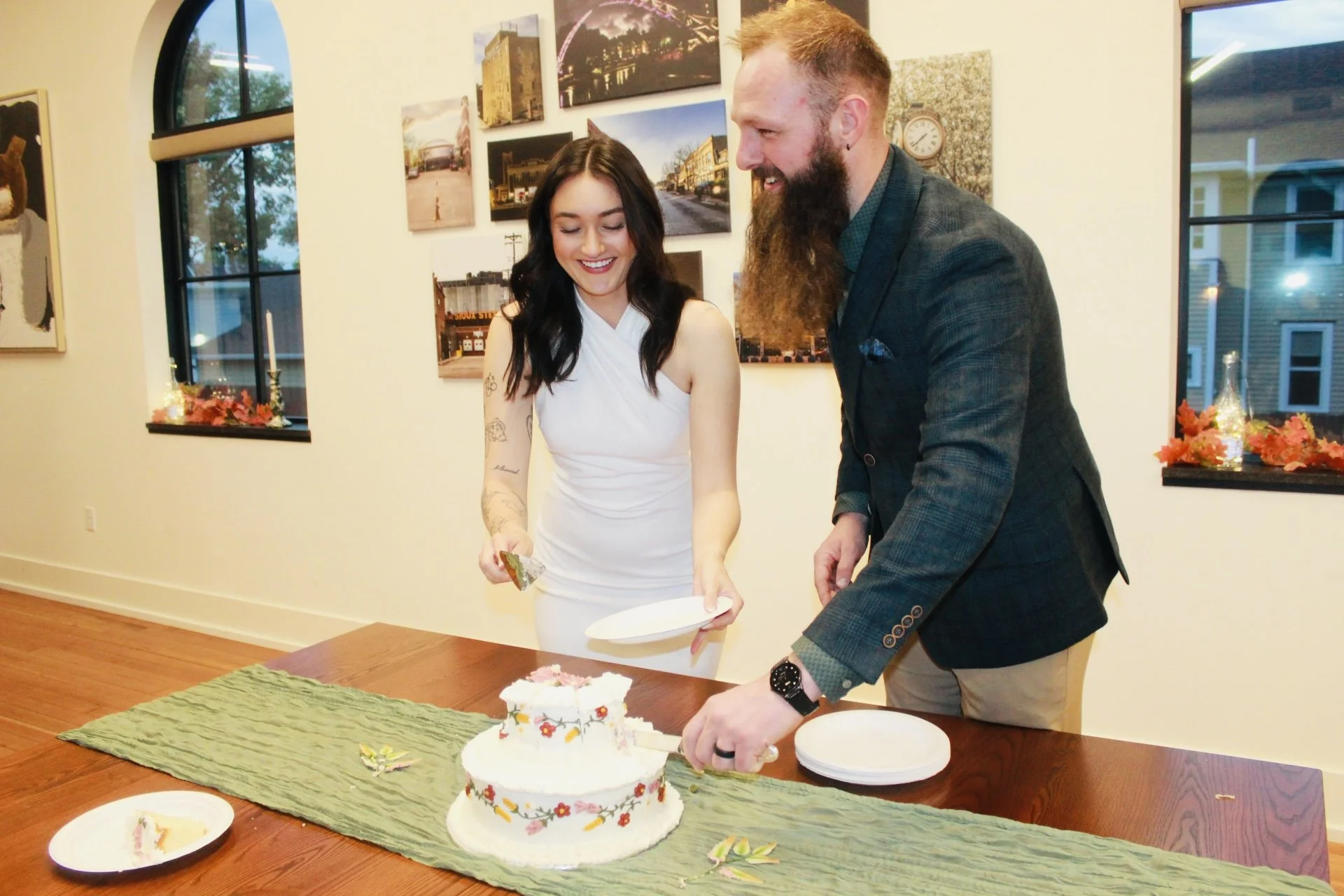 A woman in a white dress and a man in a dark blazer are smiling as they cut a decorated birthday cake together. The table has a green table runner, and there are plates, some with cake slices. The background shows walls decorated with framed artwork 