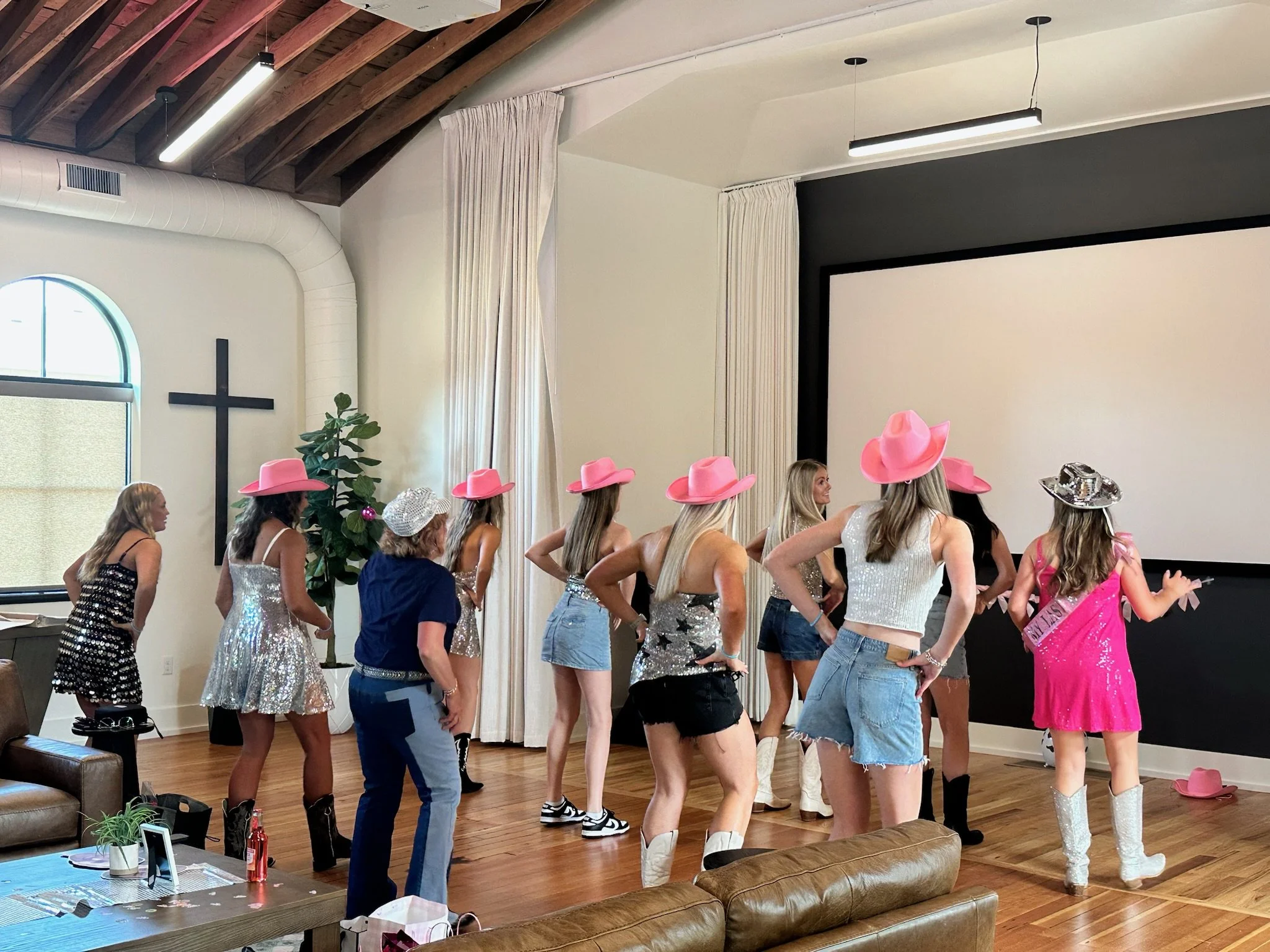 A group of women wearing cowboy hats and casual clothes standing in a line inside a room with a wooden ceiling, a cross on the wall, and a projector screen.