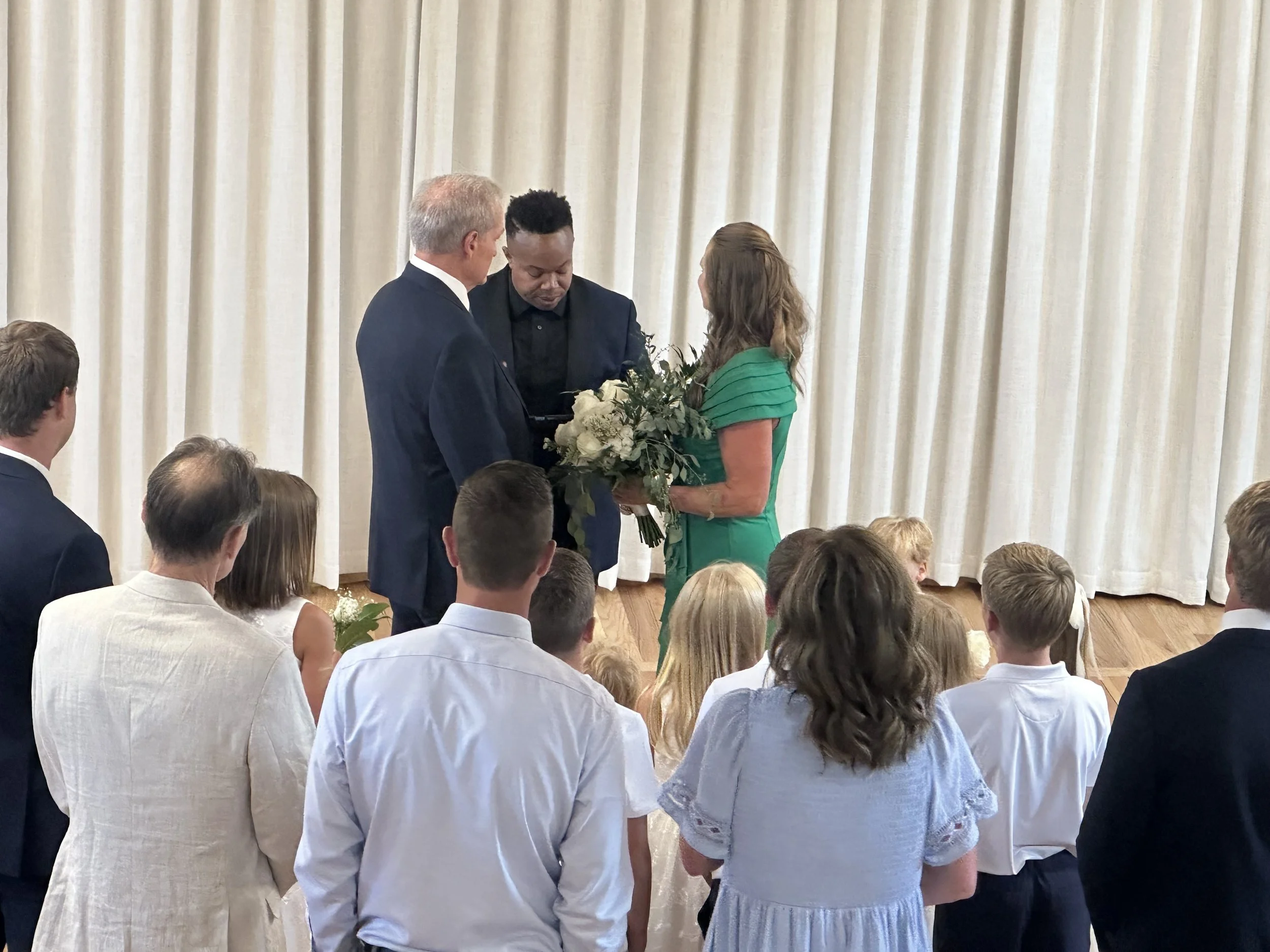 A group of people attending a formal event, with three individuals in the center exchanging a bouquet of white flowers, surrounded by others seated and facing a cream-colored curtain backdrop.