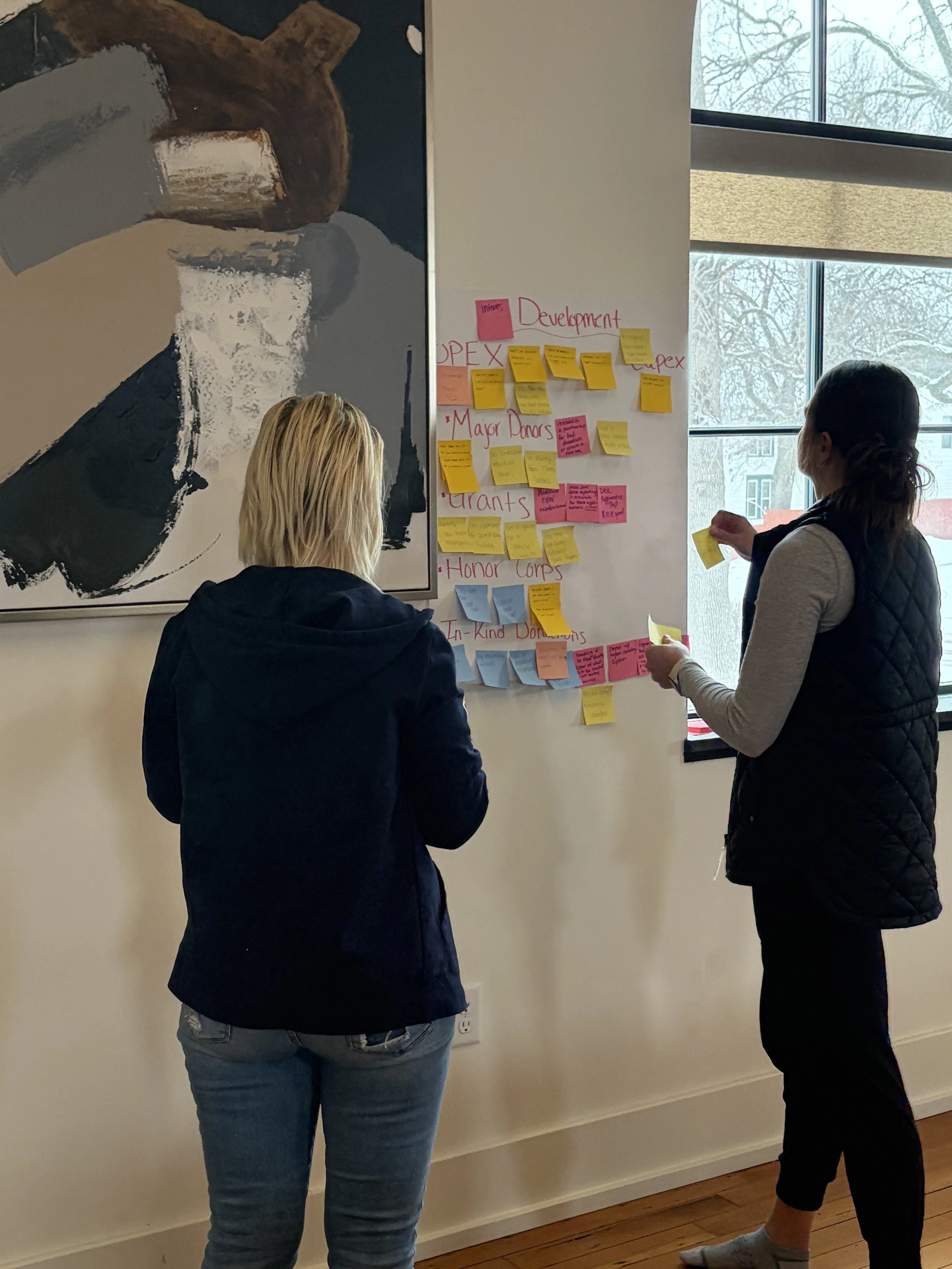 Two women are standing in front of a wall covered with colorful sticky notes, engaging in a discussion or collaboration, near a window with snow-covered trees outside.