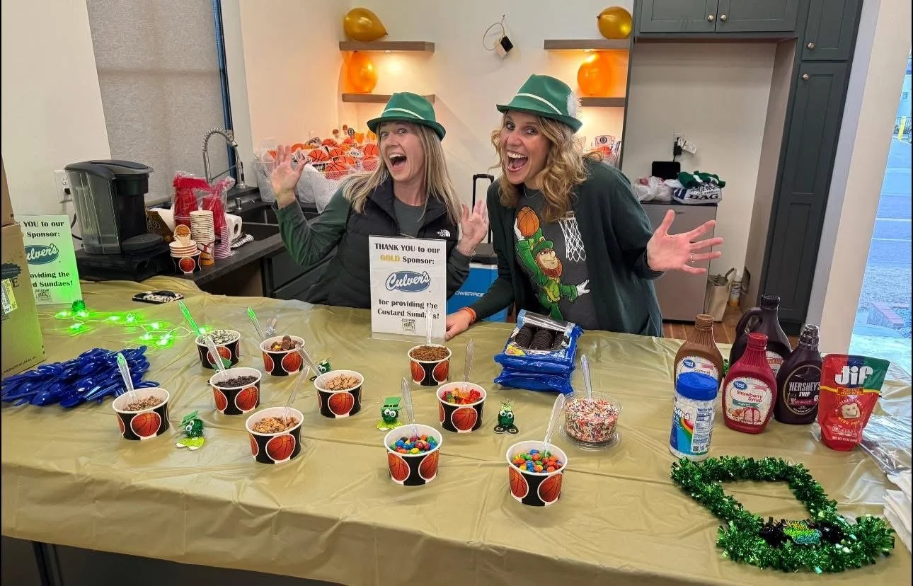 Two women cheering and smiling behind a table decorated for a basketball game celebration, wearing green hats. The table has cups with desserts, candies, and festive decorations, with a sign recognizing Culver's as a sponsor.