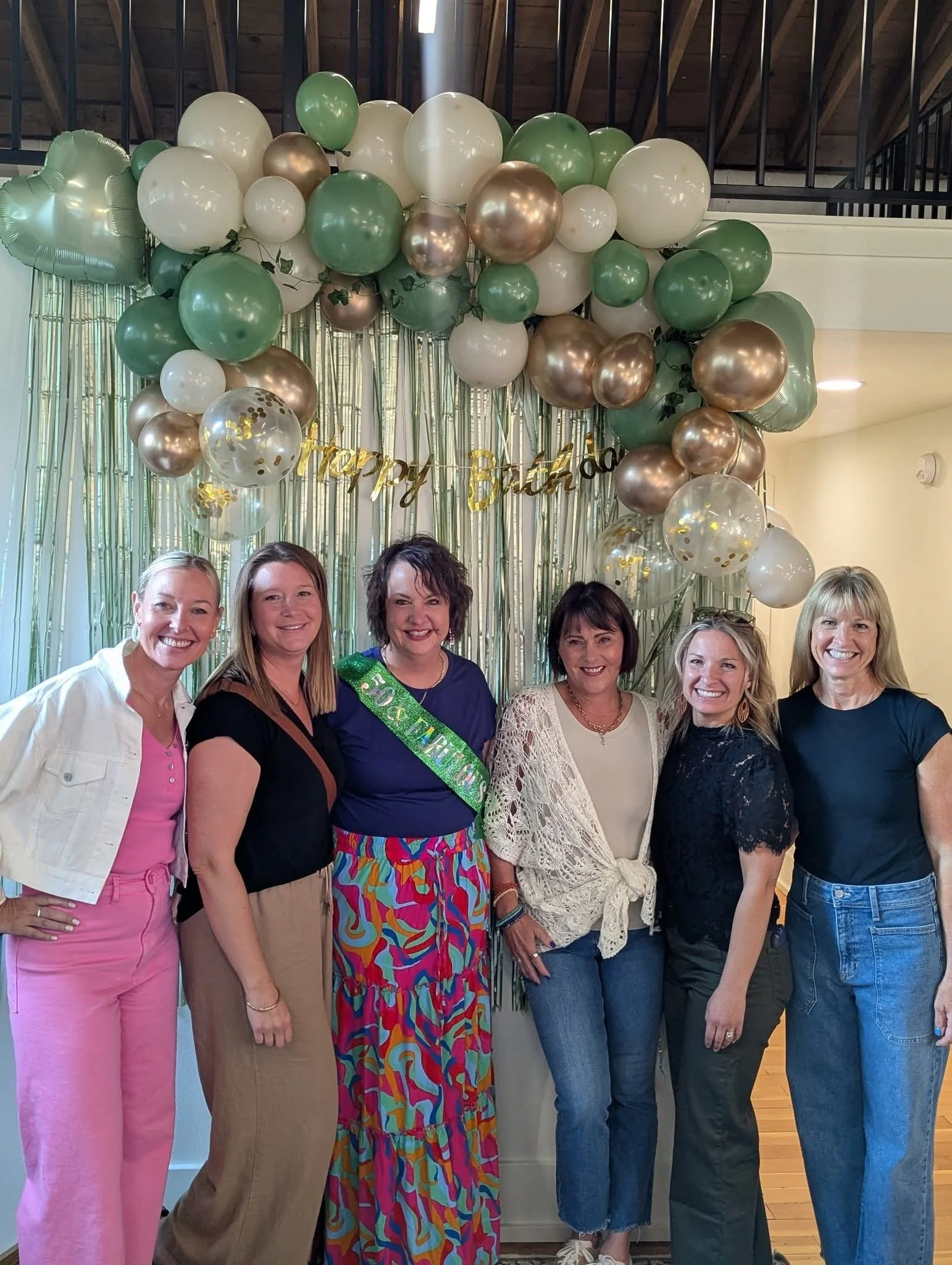 Group of six women standing together at a birthday celebration with gold, white, and green balloons and a "Happy Birthday" banner in the background.