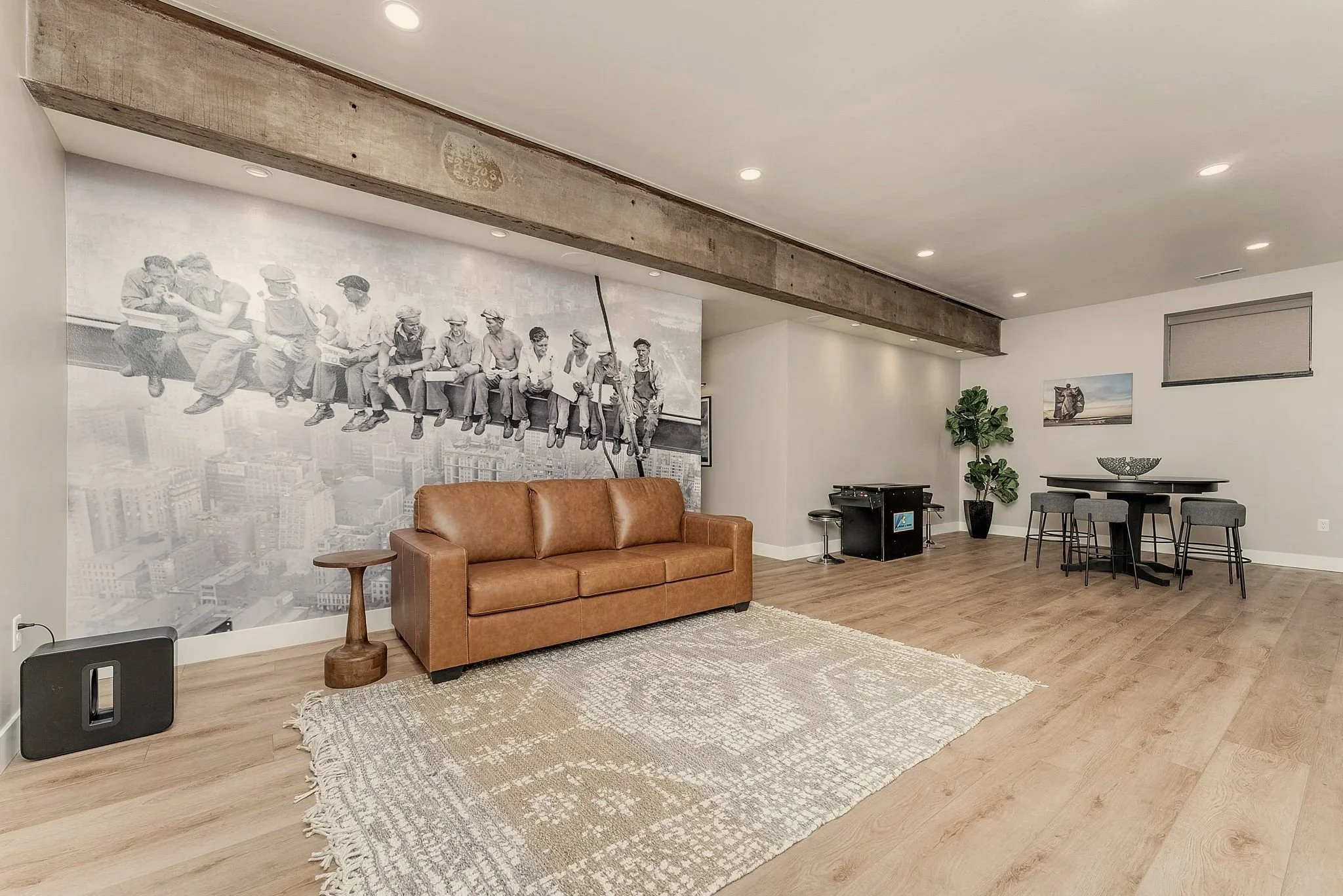 Living room with a brown leather sofa, a wooden side table, a large black-and-white photo of vintage workers on the wall, a gray rug, a potted plant, and a dining area with a round table and chairs.