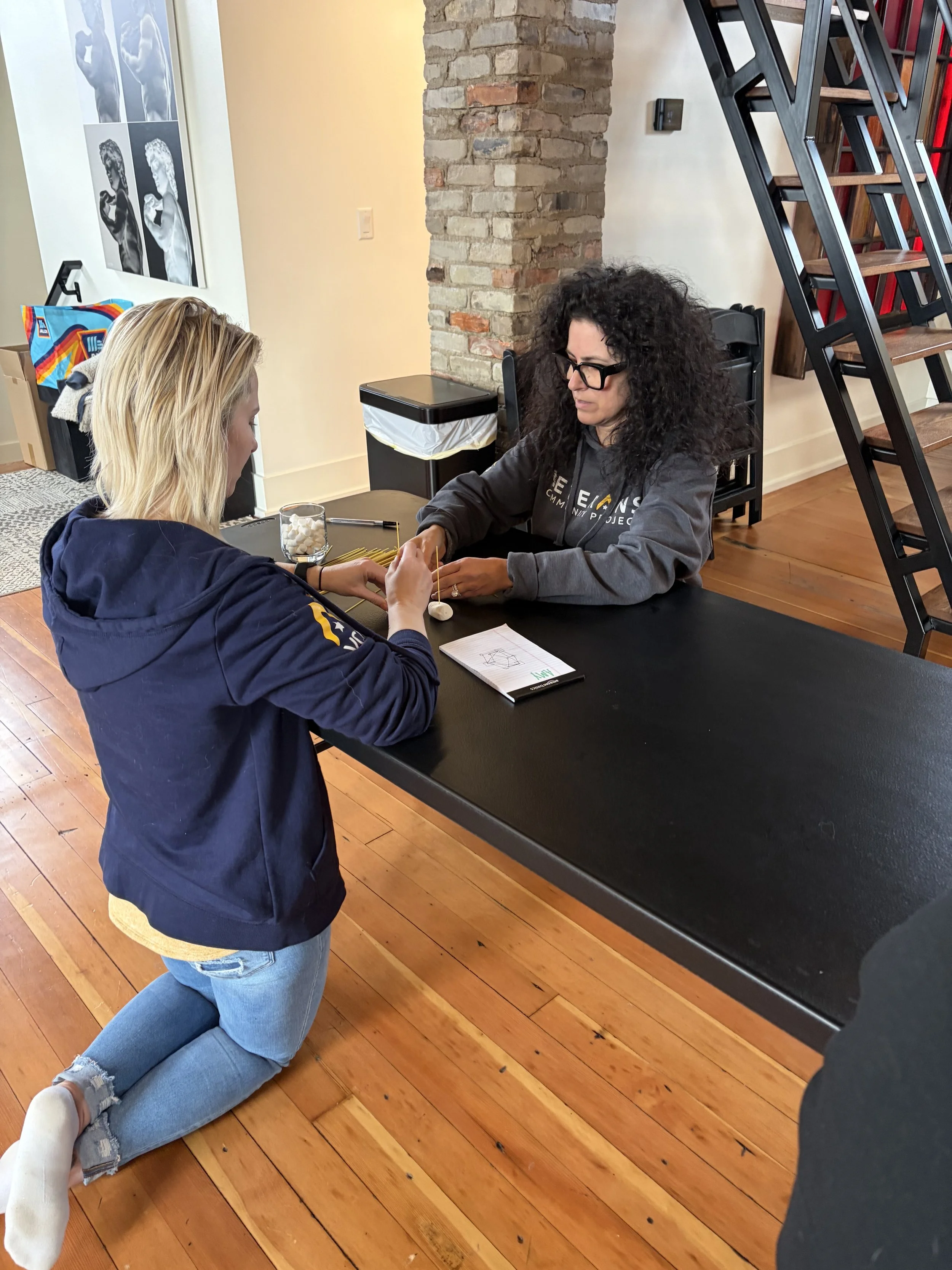 A woman kneeling on wooden floor applying a nail to a woman's finger, who is sitting at a table with a notebook, pens, a glass of white candies, and other items in a cozy indoor space with brick wall and staircase.