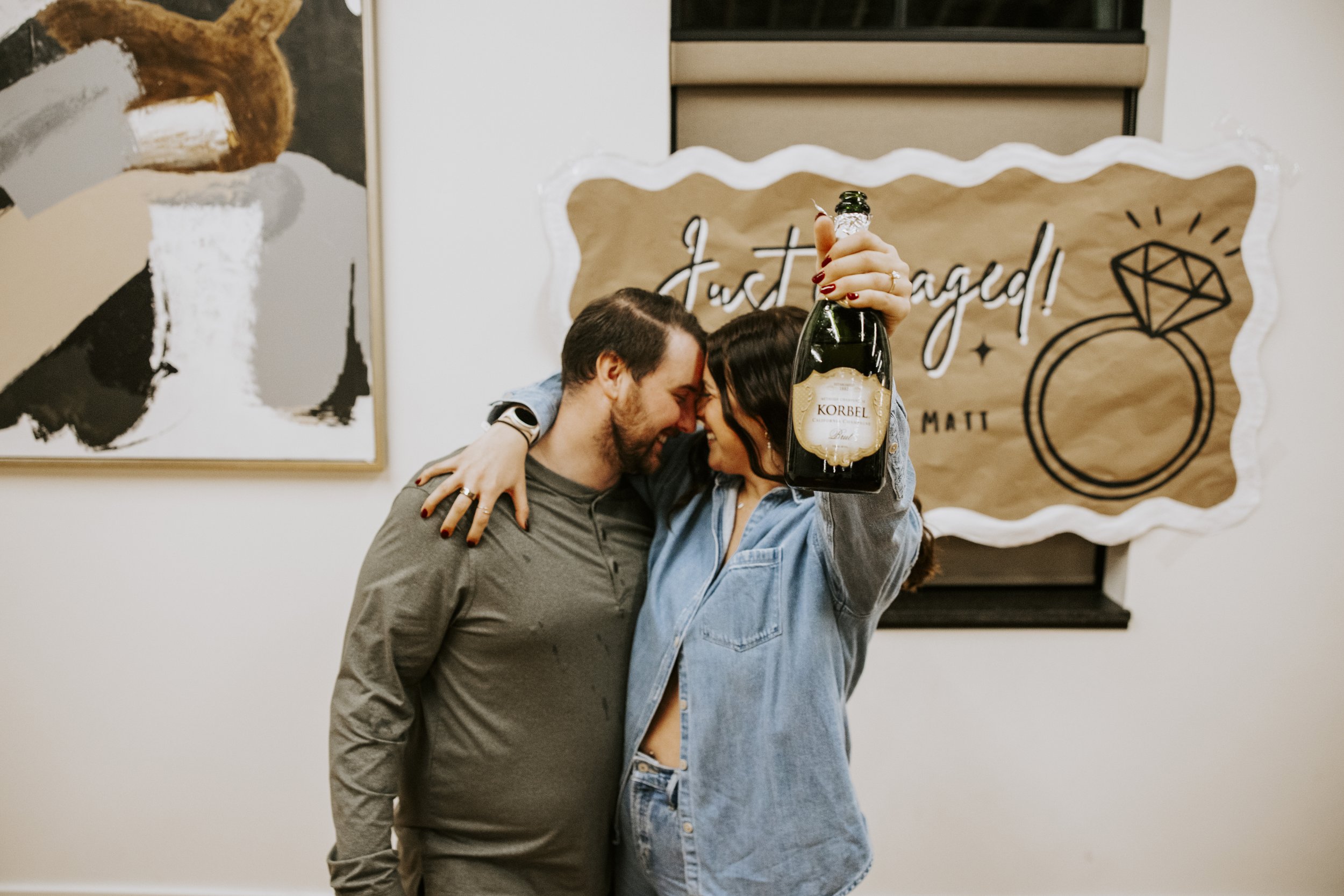A couple sharing an embrace, one holding a champagne bottle, in front of a decorated wall with a sign saying 'Just Engaged!' and a diamond ring drawing.