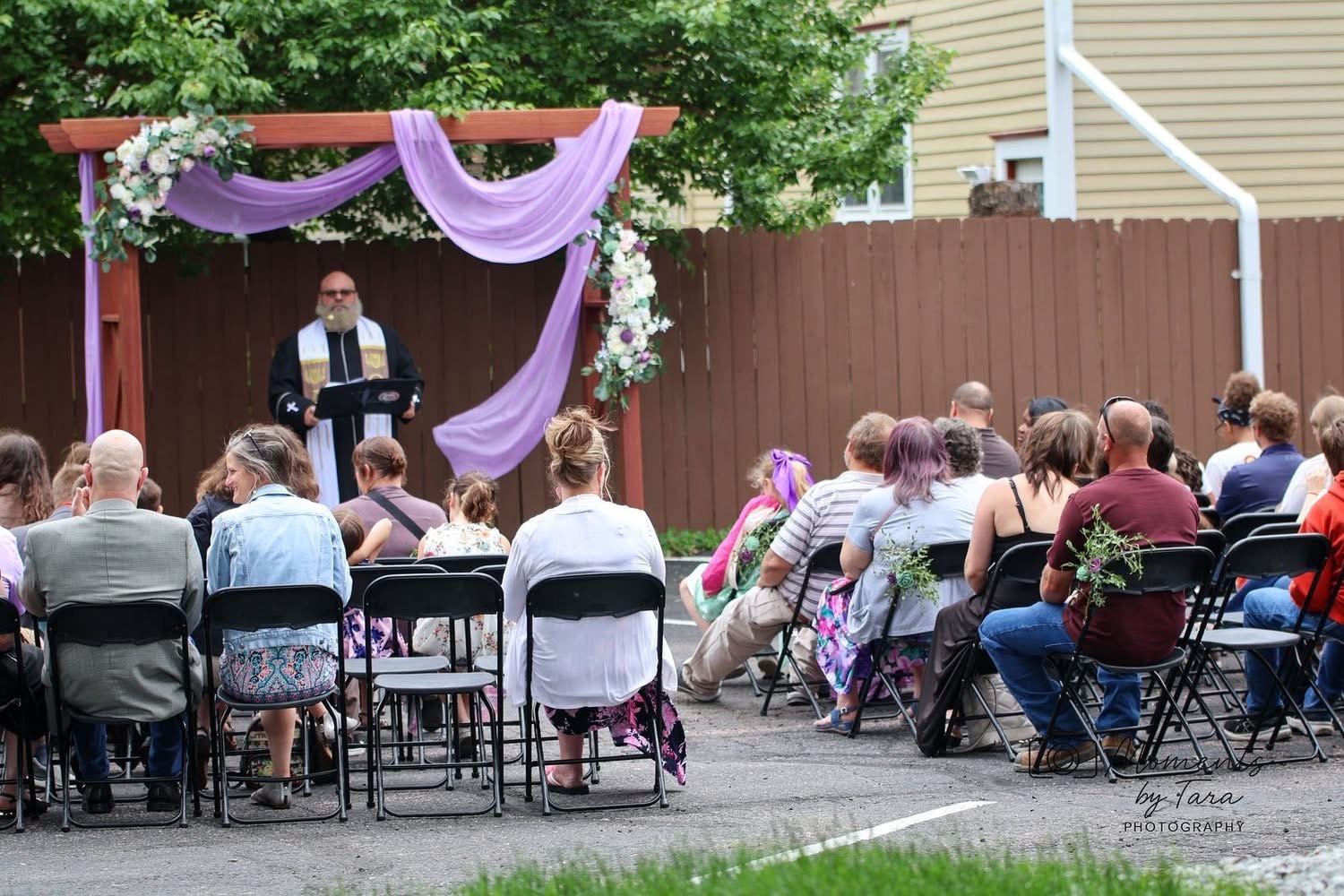 An outdoor wedding ceremony with people seated in rows, facing an altar decorated with purple drapes and flowers. A man in religious robes stands at a podium.