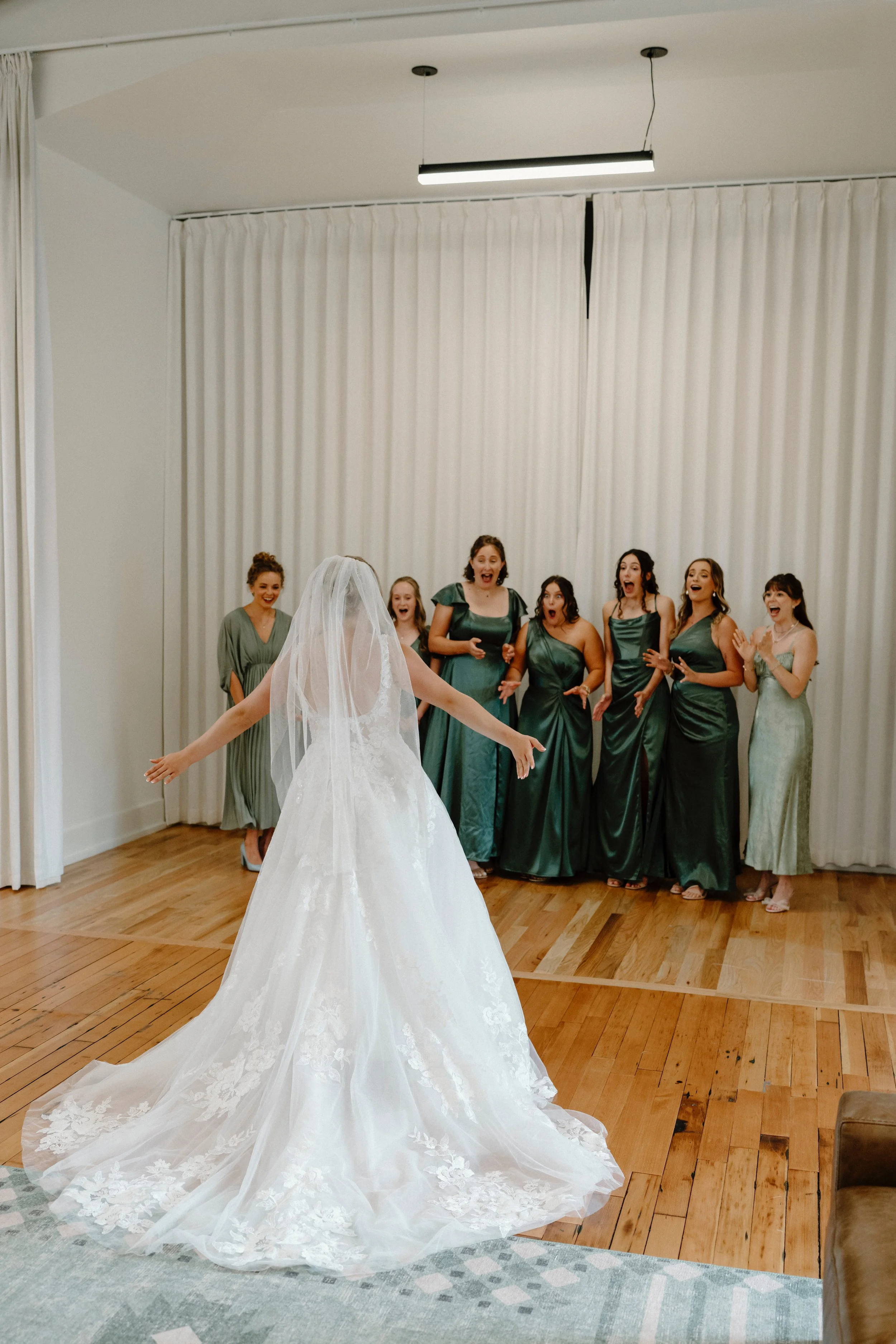 A bride in a white wedding gown and veil stands with arms outstretched in front of a group of seven women in matching green dresses, who are reacting with surprise and joy in a room with white curtains and wooden floor.