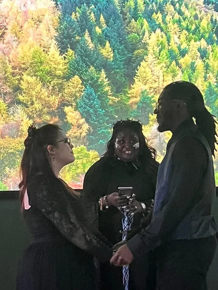 A couple is holding hands and facing each other during their wedding ceremony, with a woman officiant in the background. The couple is dressed in dark clothing, and the woman is wearing glasses. The officiant is smiling, holding a phone, and dressed 