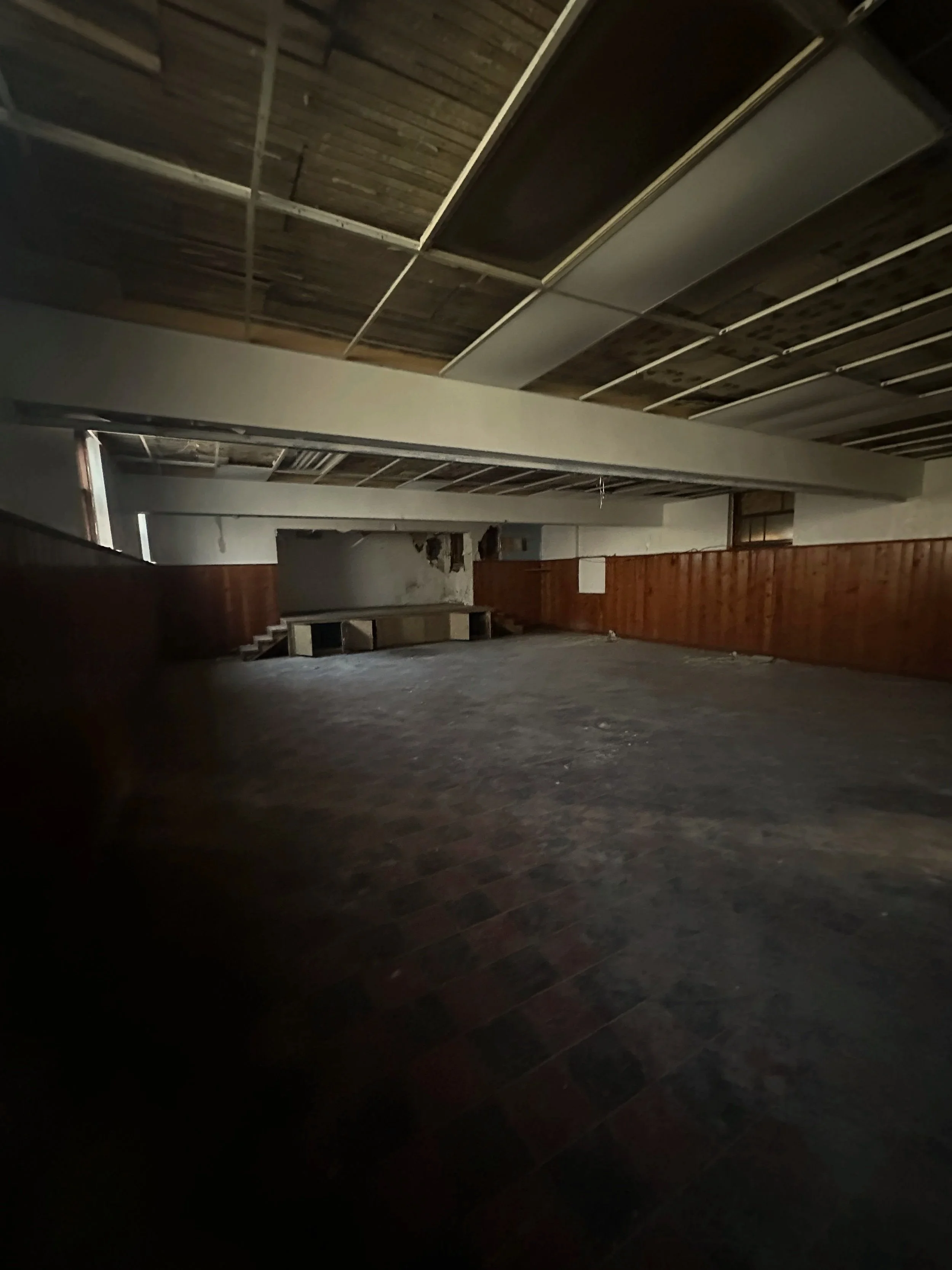 A spacious, empty room with a partially exposed wooden ceiling and dark wood flooring. The walls are a combination of white and wood paneling, with some areas showing damage or missing sections.
